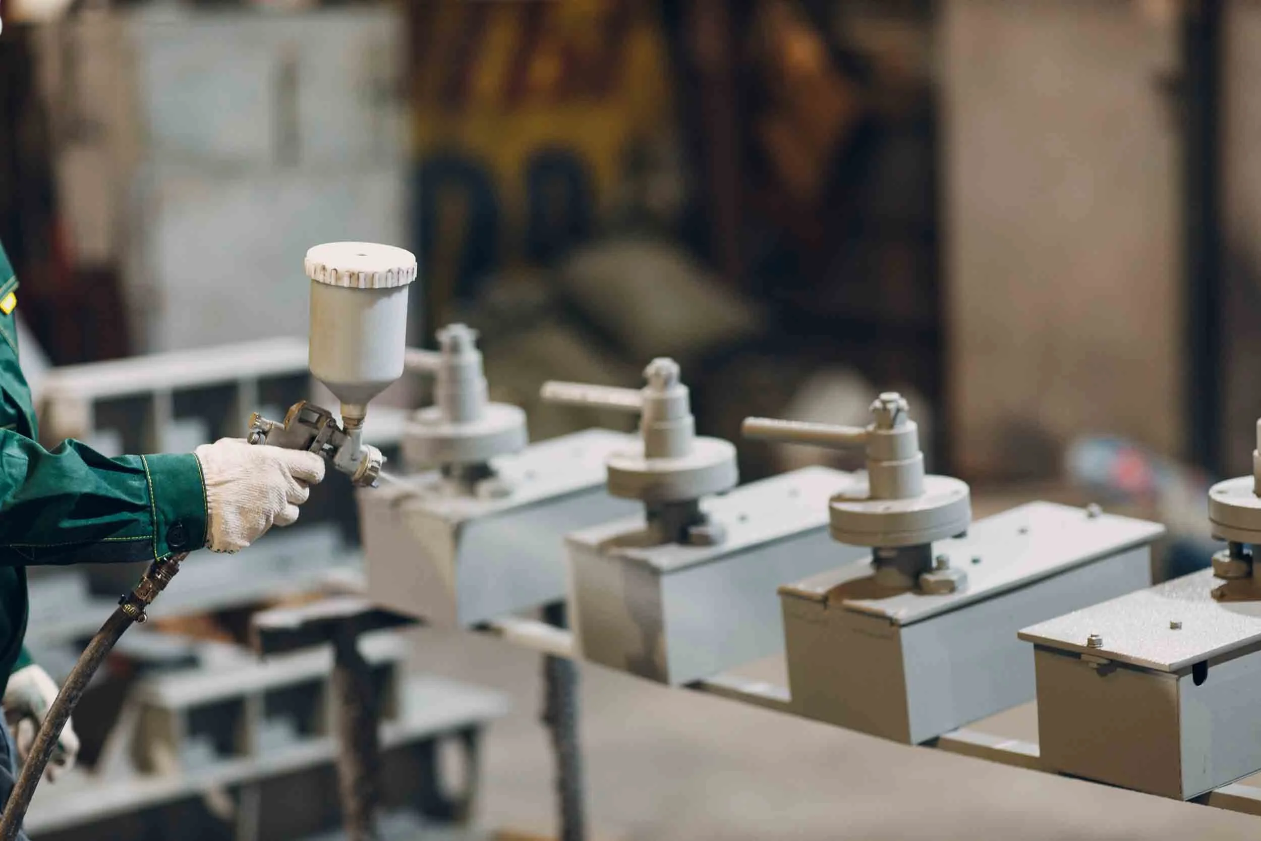 A worker wearing gloves using a spray gun to apply paint or coating to industrial equipment or components in a manufacturing or assembly setting.