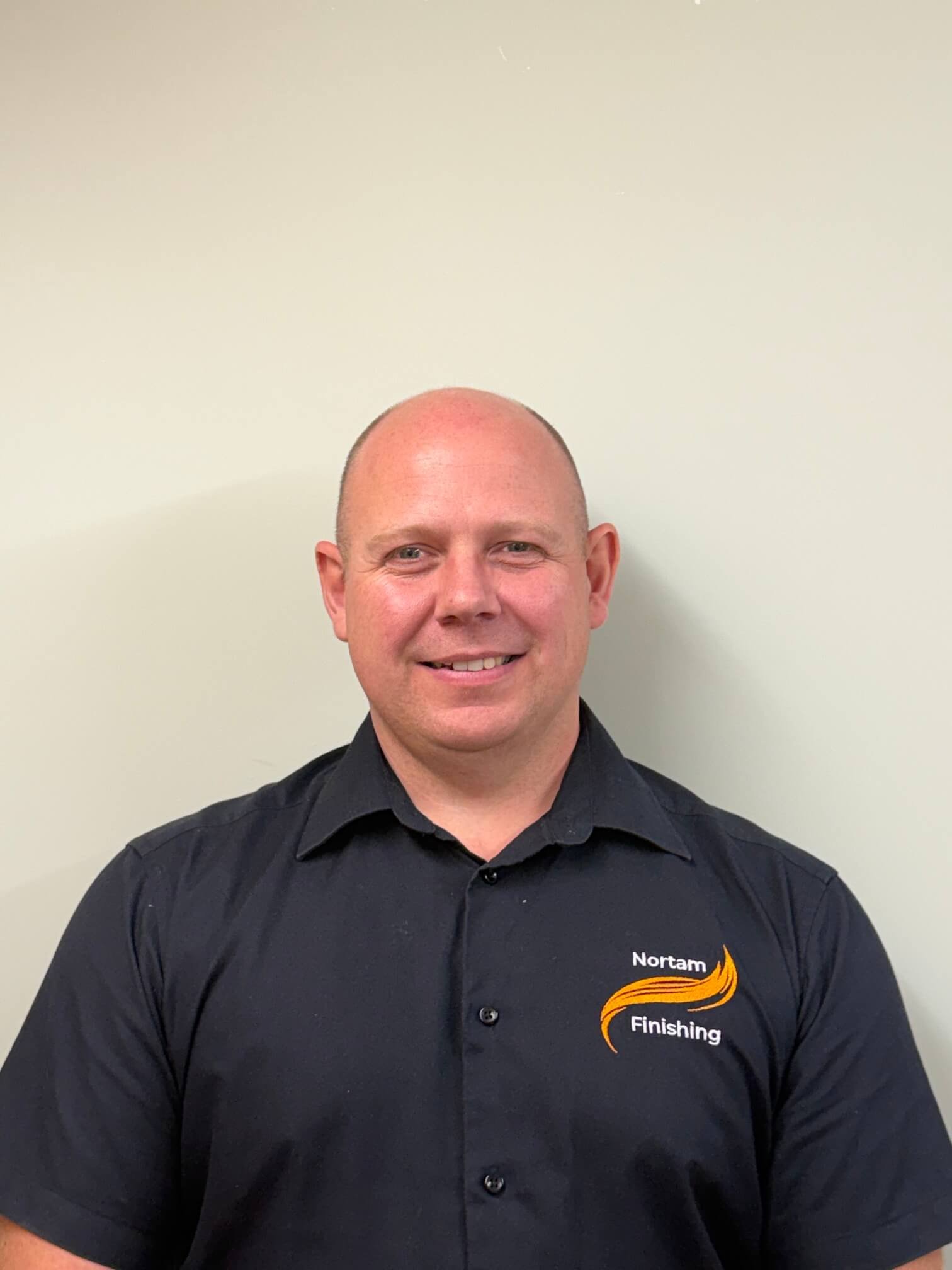 A man with a big smile, wearing a black shirt with a logo that reads 'Nortam Finishing' on the chest, stands against a plain light-colored wall.