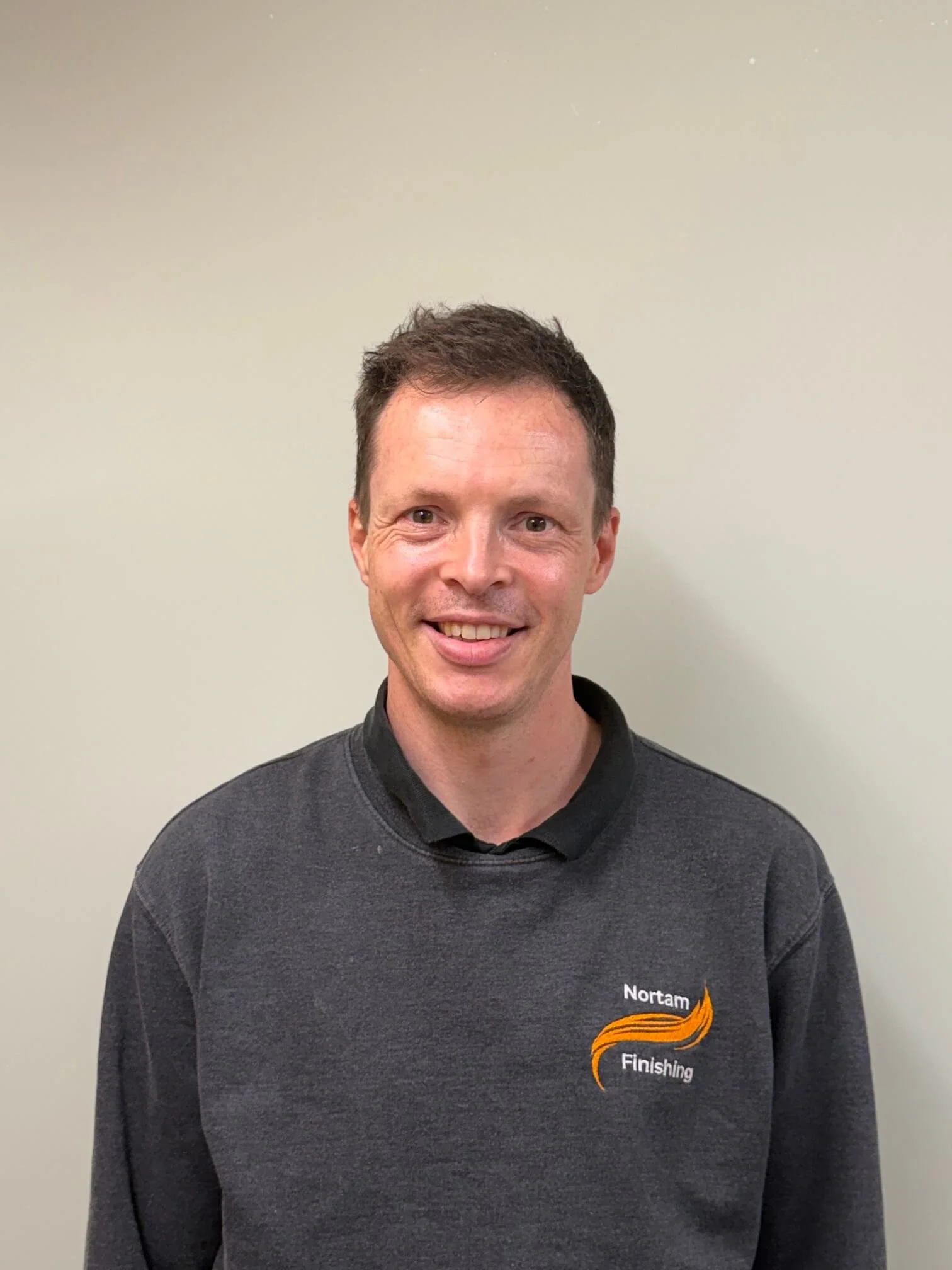 A smiling man with short brown hair wearing a dark gray sweatshirt with the logo 'Nortam Finishing' and a background of a plain light-colored wall.