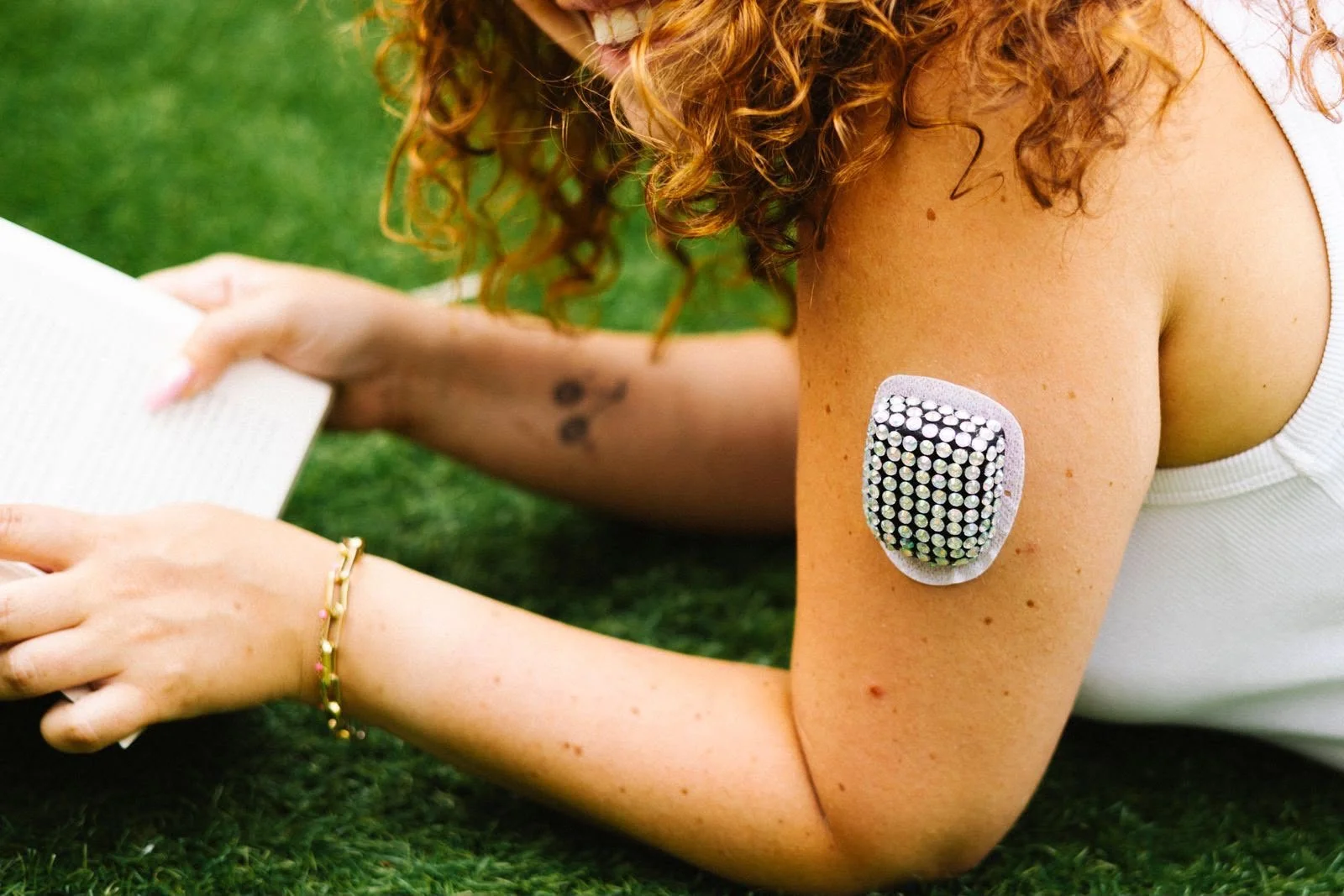 A woman with curly red hair lying on grass, wearing a white top, with a decorative rhinestone patch on her shoulder, a gold bracelet, and a small tattoo of three black circles on her arm, reading a book.