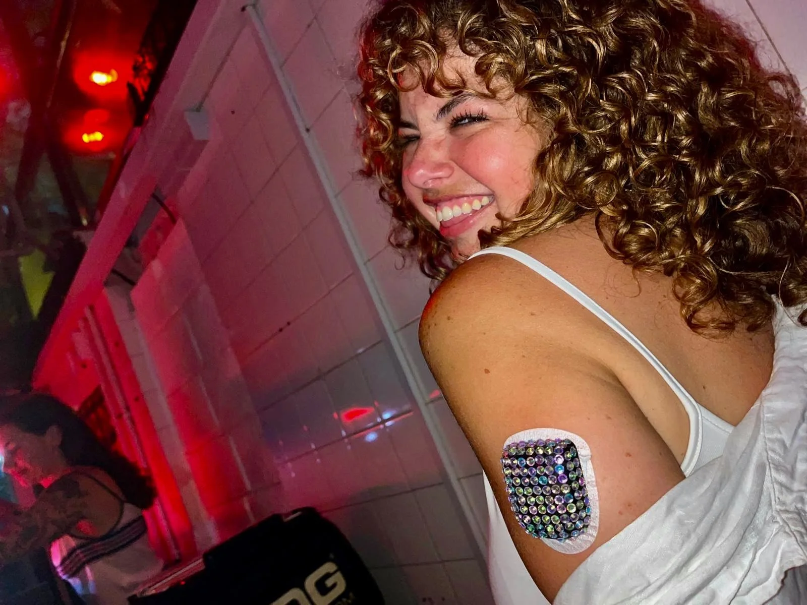 A woman with curly blonde hair smiling at a party. She has a decorative bandage on her upper arm that is covered in rhinestones or sequins. The background features a tiled ceiling and red lighting.