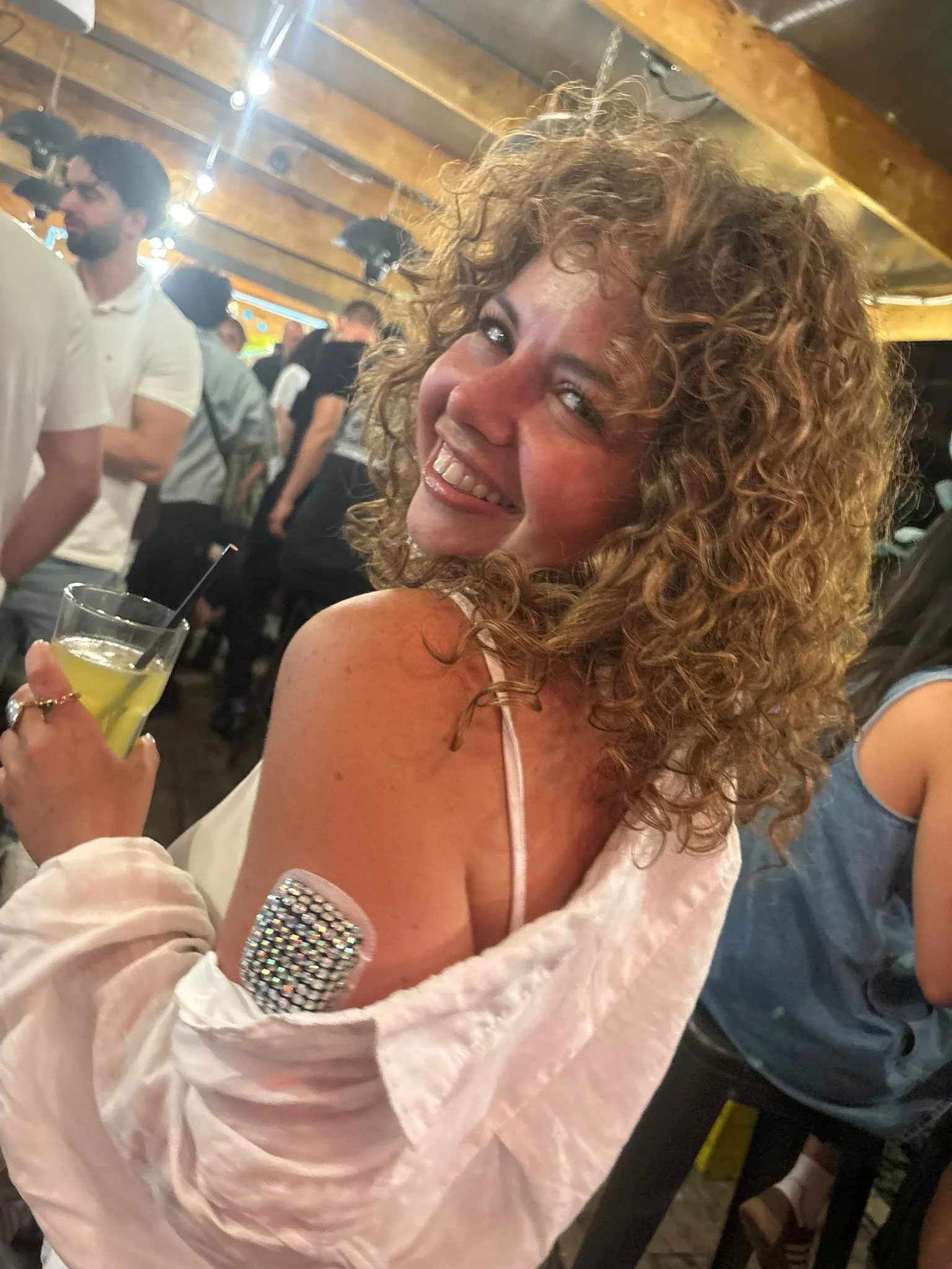 A woman with curly hair smiling and holding a drink in a crowded indoor setting with people and wooden beams overhead.