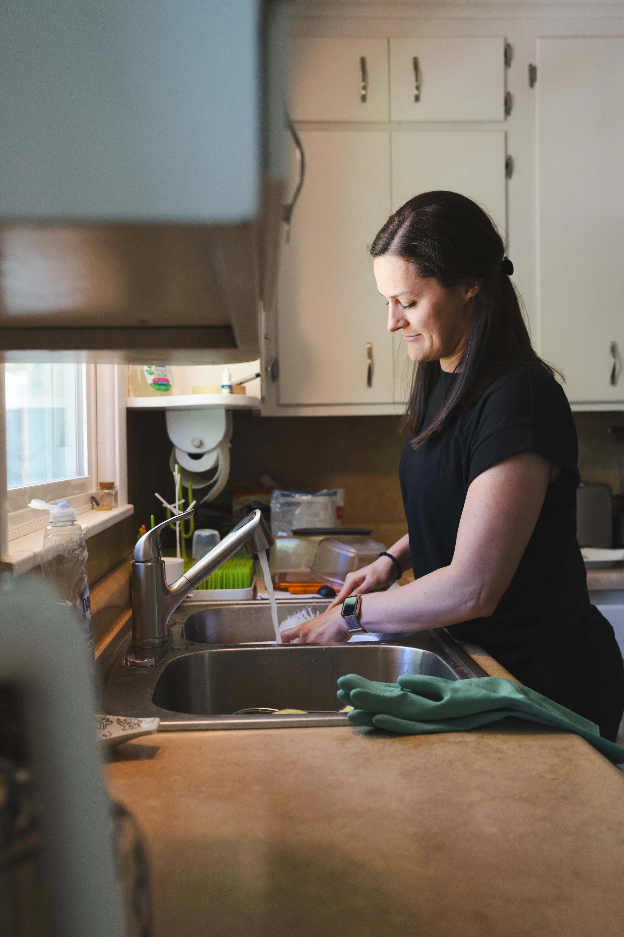 A woman with long dark hair is washing dishes at a kitchen sink, wearing a black shirt and a smartwatch. The kitchen has white cabinets, and there are various kitchen items around, including a green dish drying mat and a pair of green gloves on the counter.