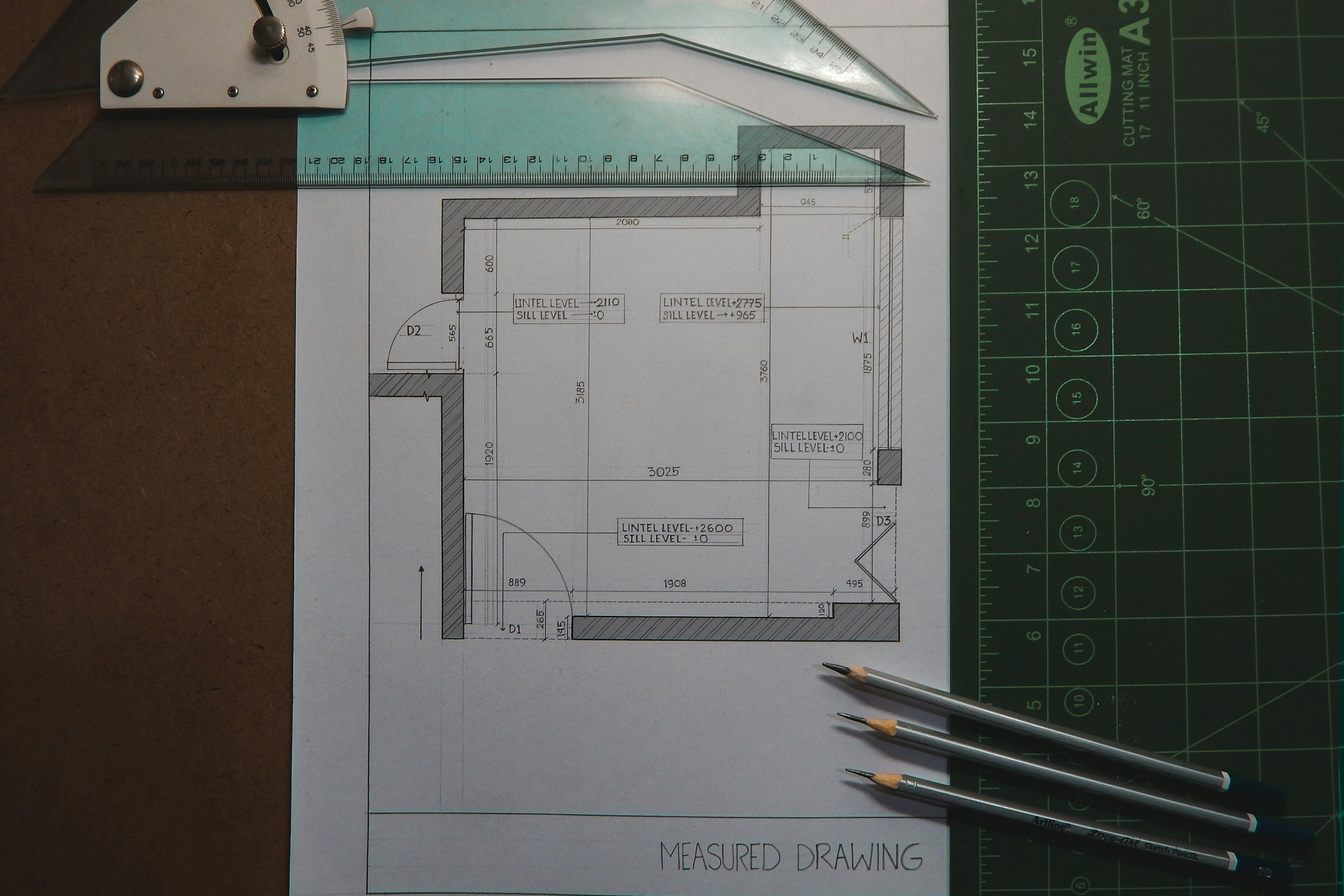 A measured architectural drawing of a house floor plan on paper, along with a green cutting mat, three precision tools, and a mechanical pencil, all used for drafting.