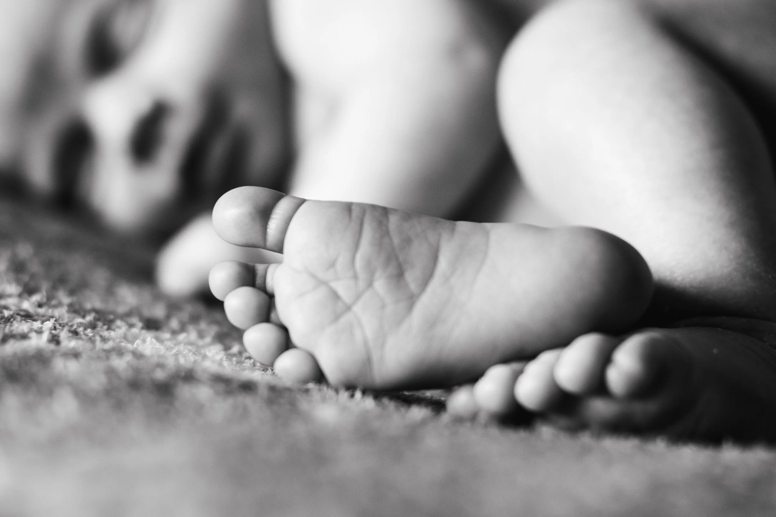 Close-up black and white photo of a baby's feet lying on a textured surface, with the background out of focus.