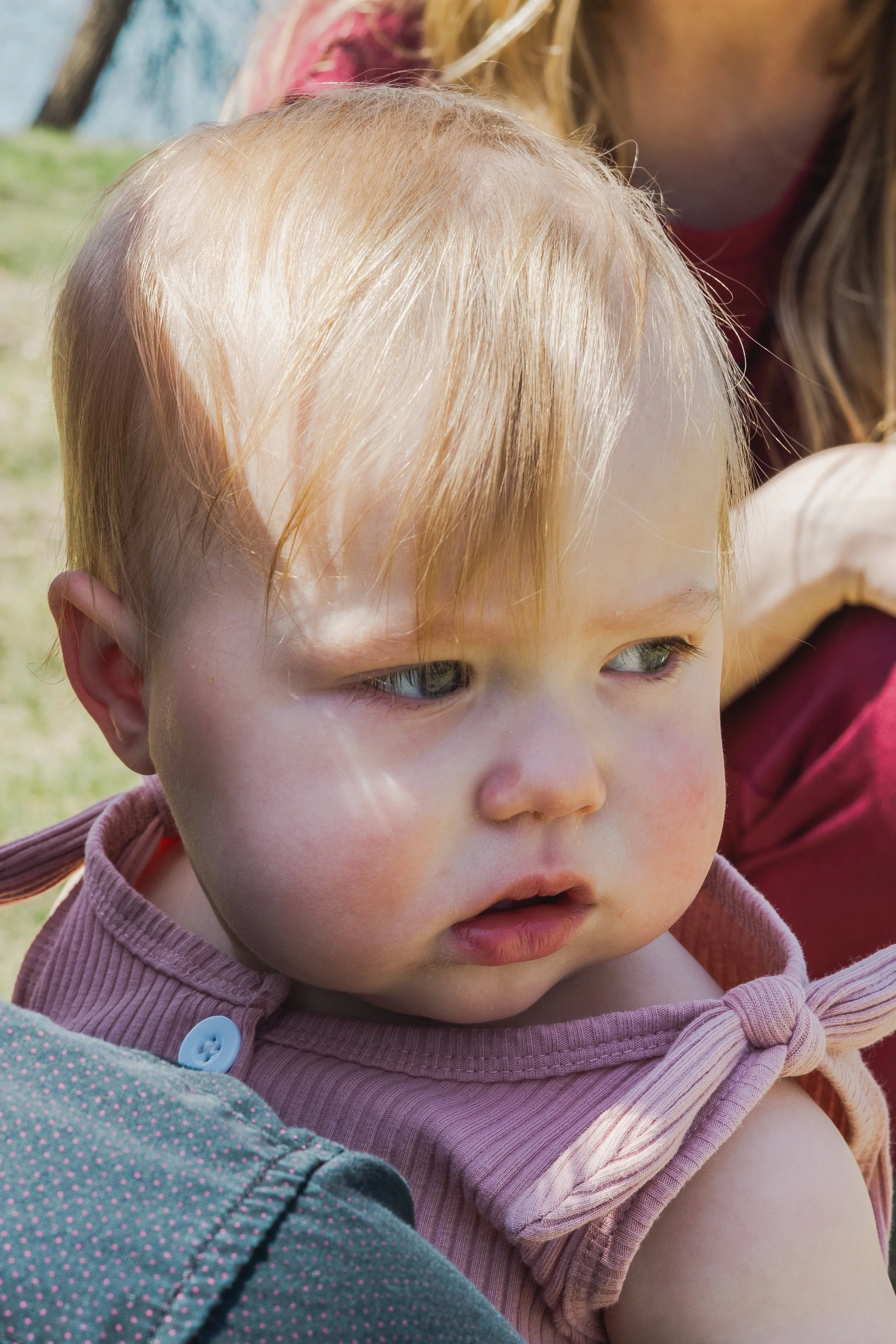Close-up of a young child with blonde hair, blue eyes, wearing a pink and gray polka-dotted shirt, outdoors with another person partially visible behind.