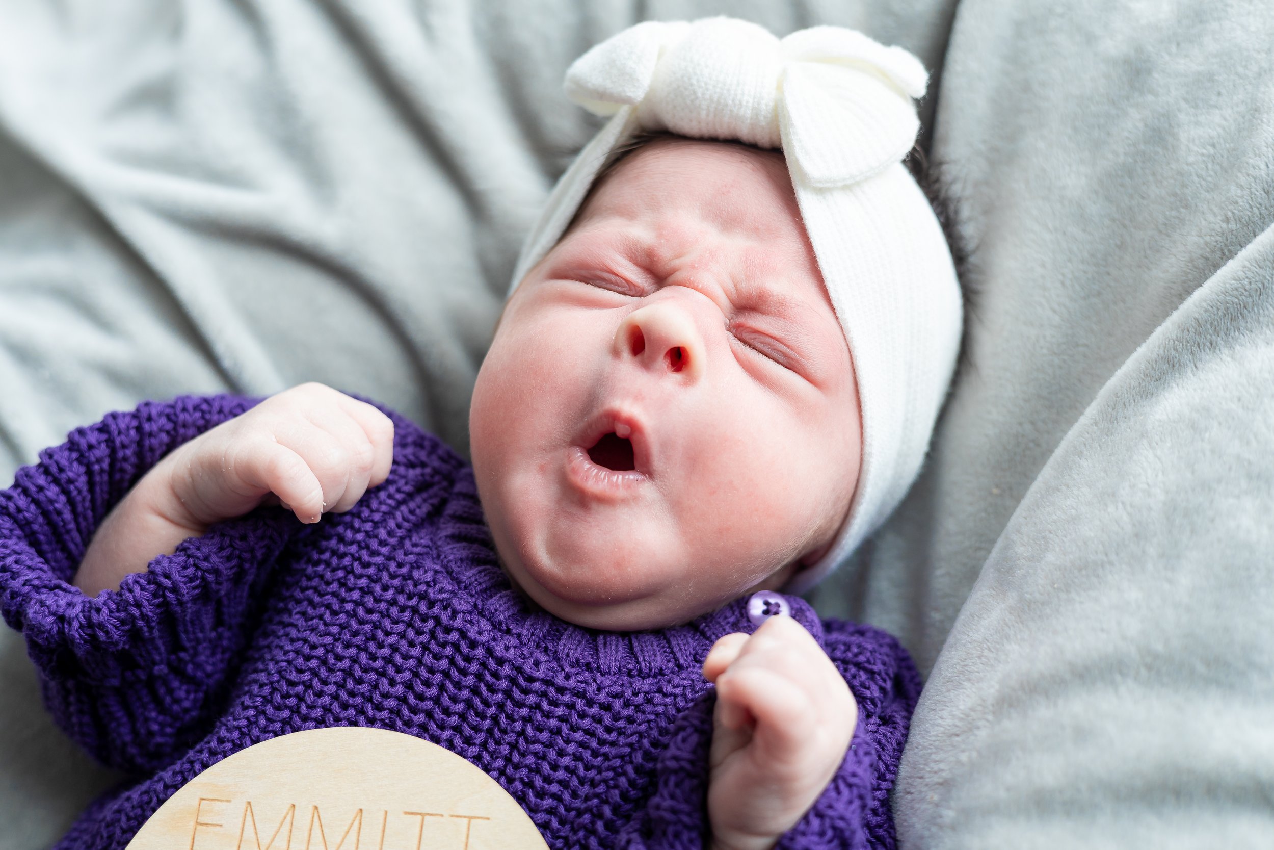 Close-up of a newborn baby girl yawning with closed eyes and mouth open, wearing a white hat with a bow and a purple knitted sweater, lying on a soft gray surface with a wooden name tag reading 'Emmitt' nearby.