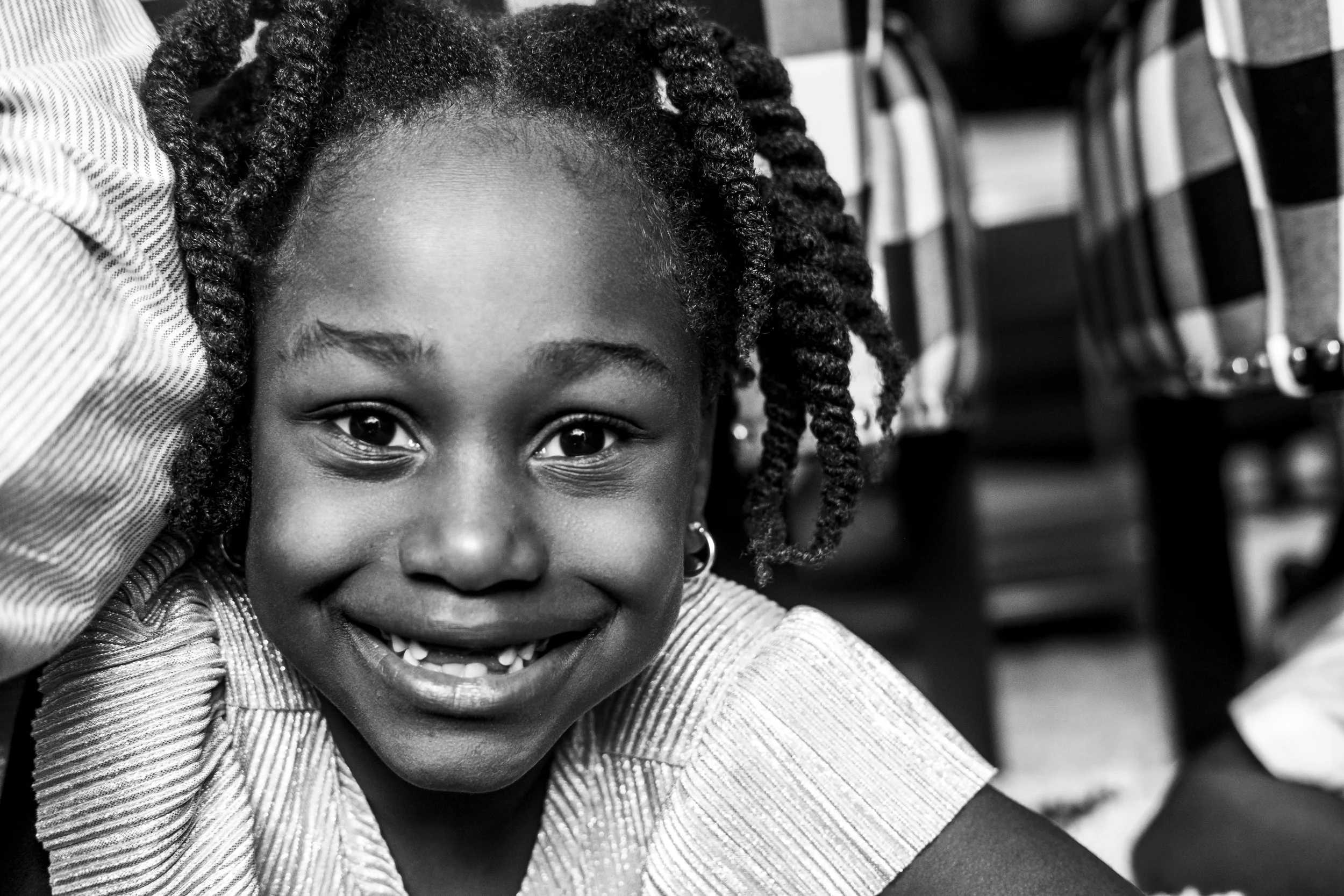 Close-up black and white photo of a smiling young girl with braided hair looking at the camera, with blurred chairs in the background.