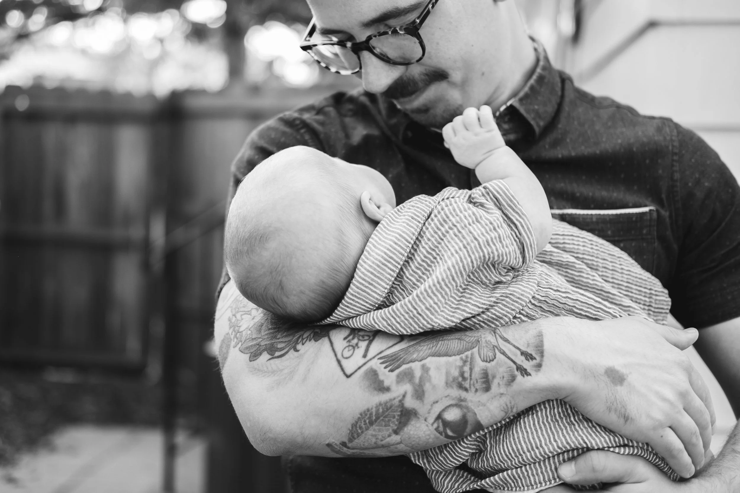 A man with tattoos and glasses holding a baby who is touching his face in an outdoor setting.