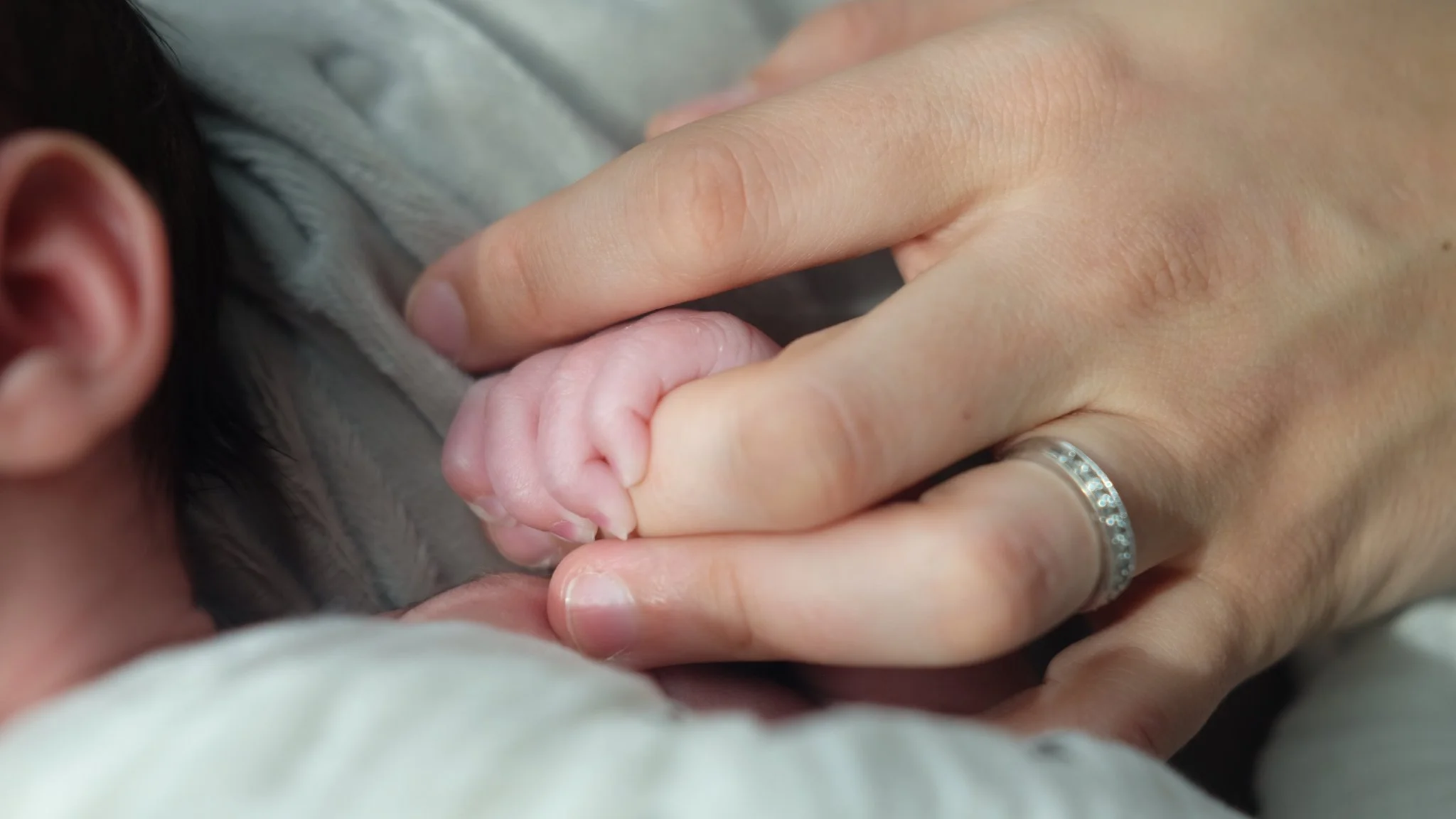 Close-up of a person's hand holding a newborn baby's tiny hand, showing a ring on the adult's ring finger.
