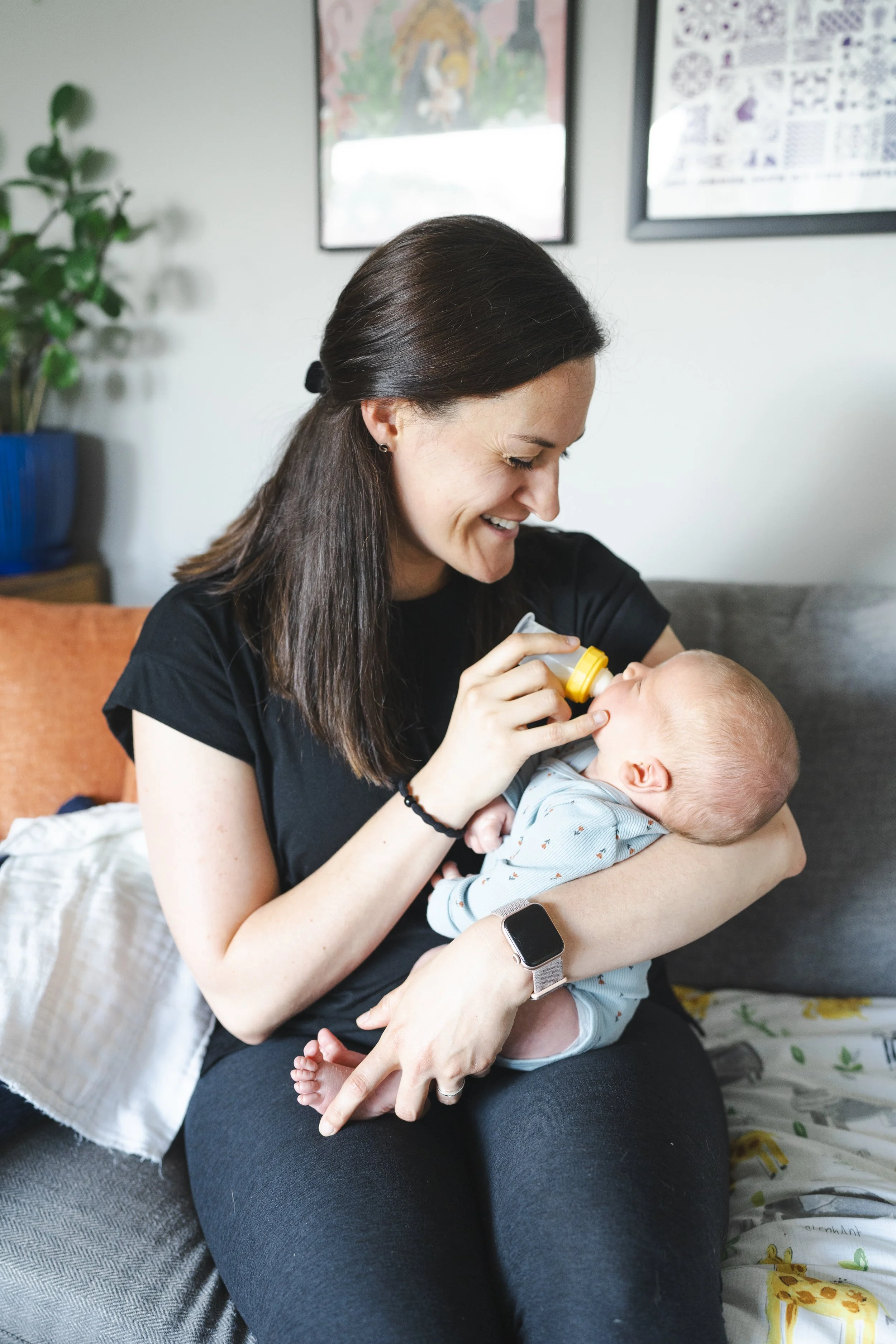 A woman with dark brown hair, wearing a black t-shirt, is sitting on a bed, holding a baby and feeding it with a bottle. The woman is smiling and looking down at the baby. In the background, there are framed pictures on the wall, a potted plant on a wooden surface, and a bed with colorful bedding.