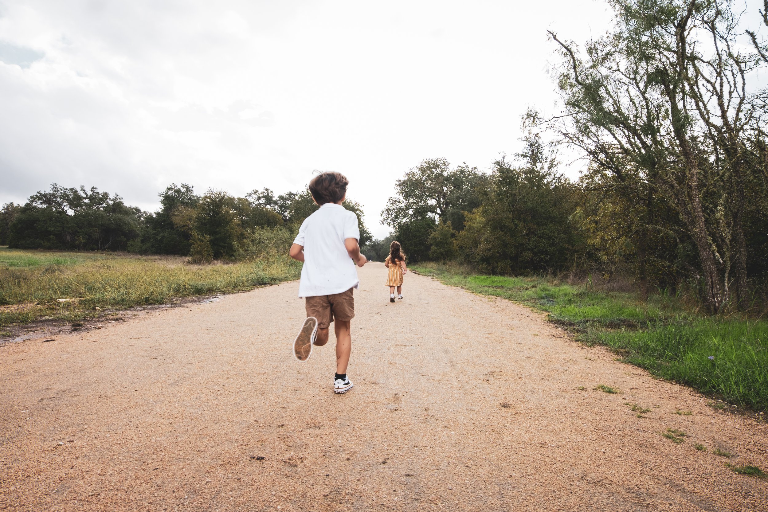 Two children running on a dirt path through a natural landscape with trees on either side, under a partly cloudy sky.