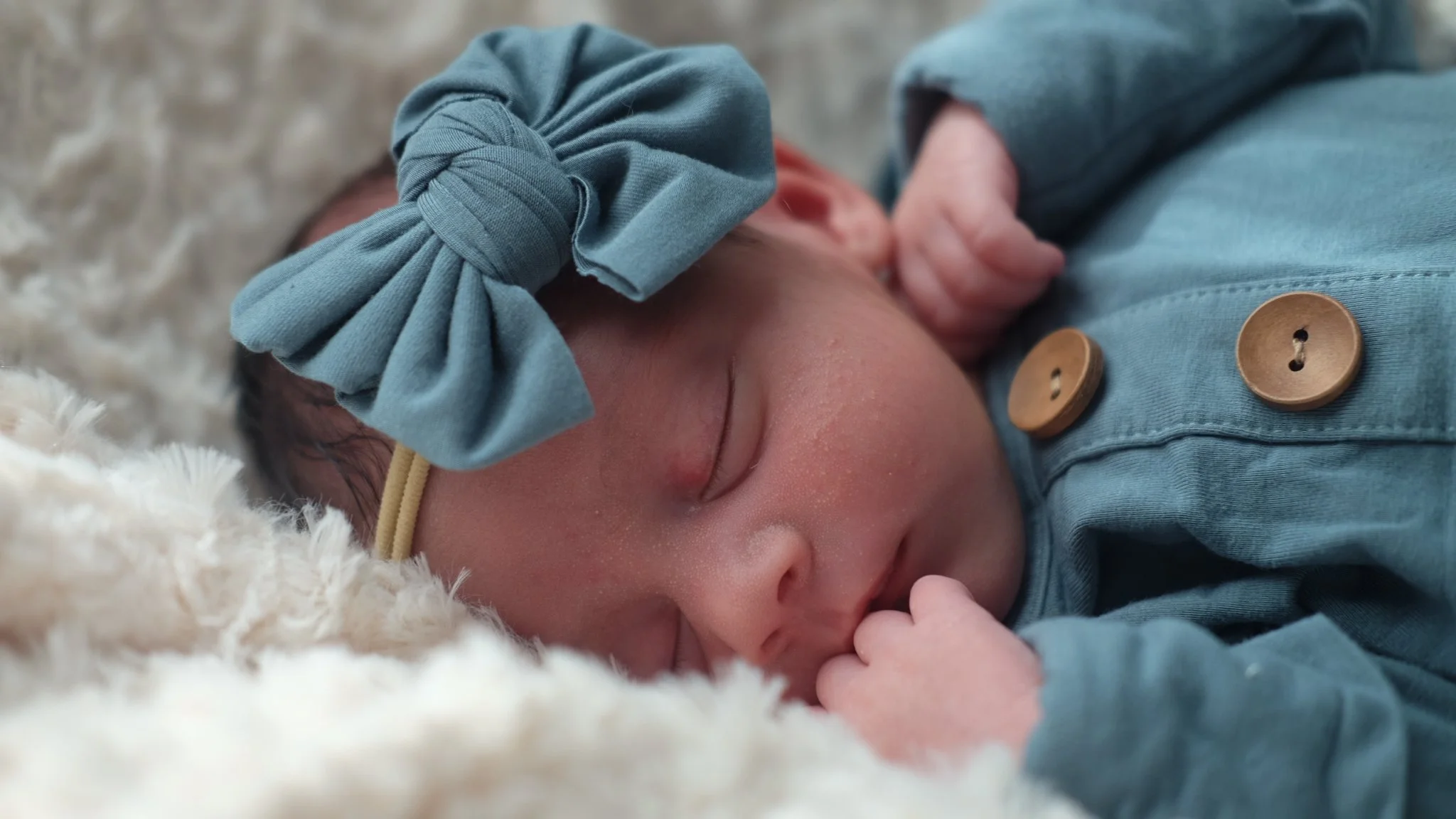Close-up of a sleeping newborn baby wearing a blue headband with a bow, dressed in a blue outfit with wooden buttons, resting on a soft white blanket.