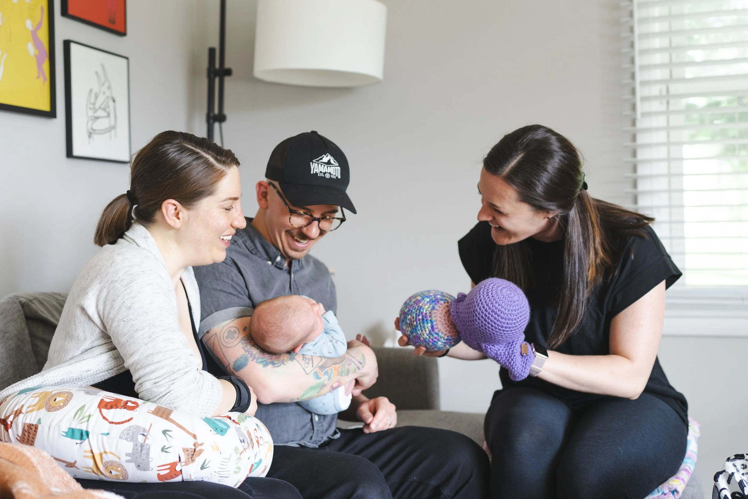 A group of people, including two women and a man, gathered around a baby and a doll. They are smiling and appear to be engaging in a playful activity together in a bright room with artwork on the walls and window blinds in the background.