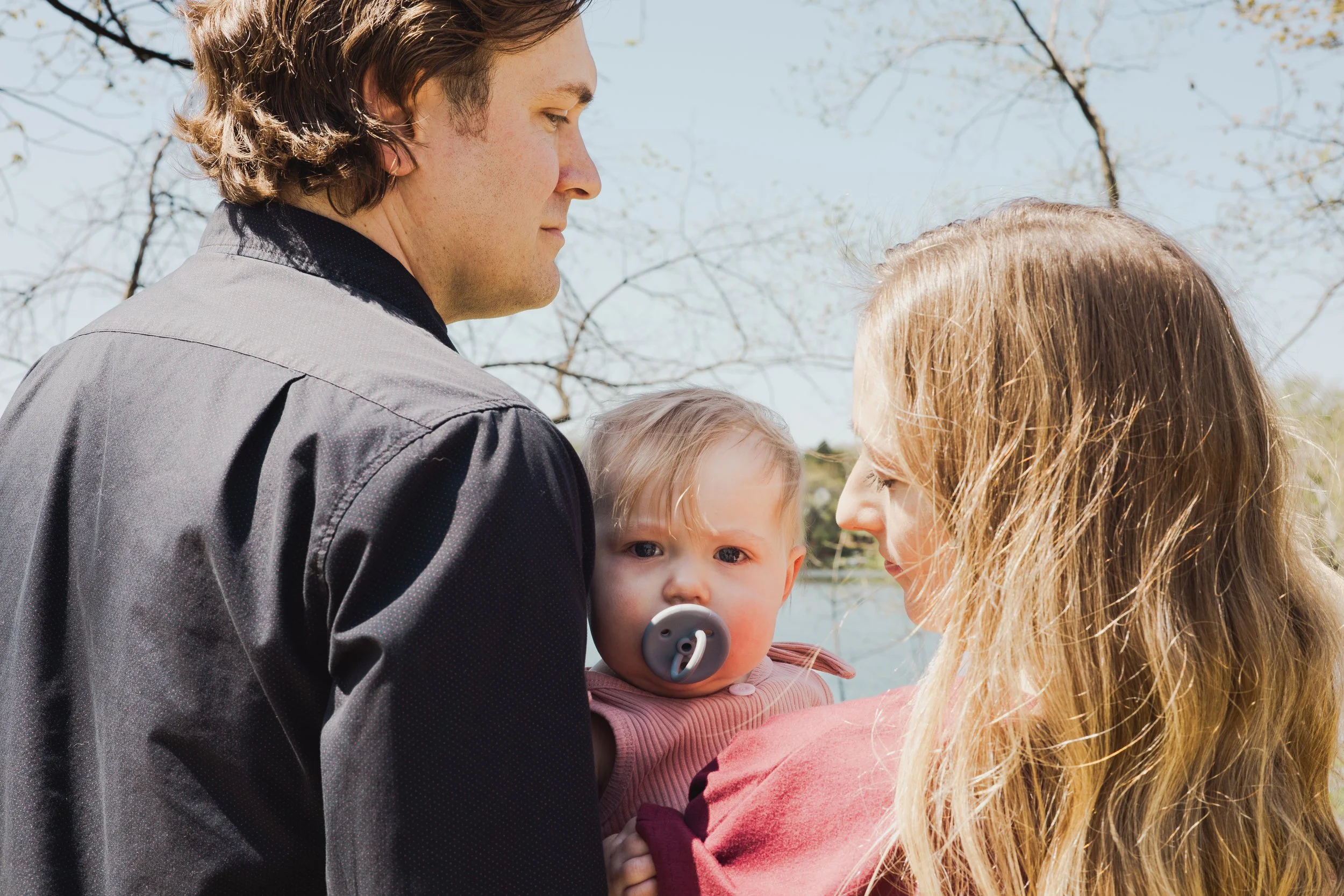 A man, woman, and child are close together outdoors, with the man on the left and the woman and child on the right. The man has brown, wavy hair and is wearing a gray jacket. The woman has long, blonde hair, and the child ha light-colored hair. They 