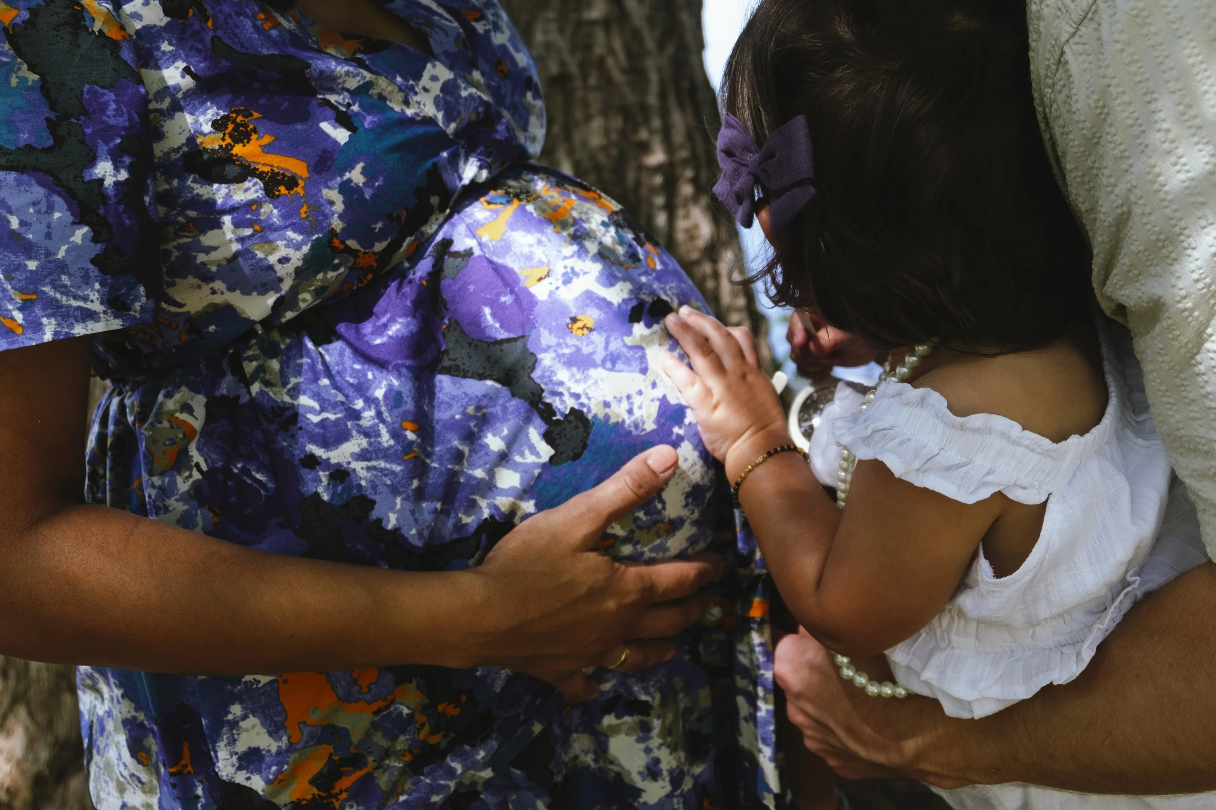 A young girl with a purple bow in her hair is holding and touching an adult person's pregnant belly. The person has on a colorful, blue, purple, black, white, and orange patterned shirt. They are outdoors, possibly near trees.