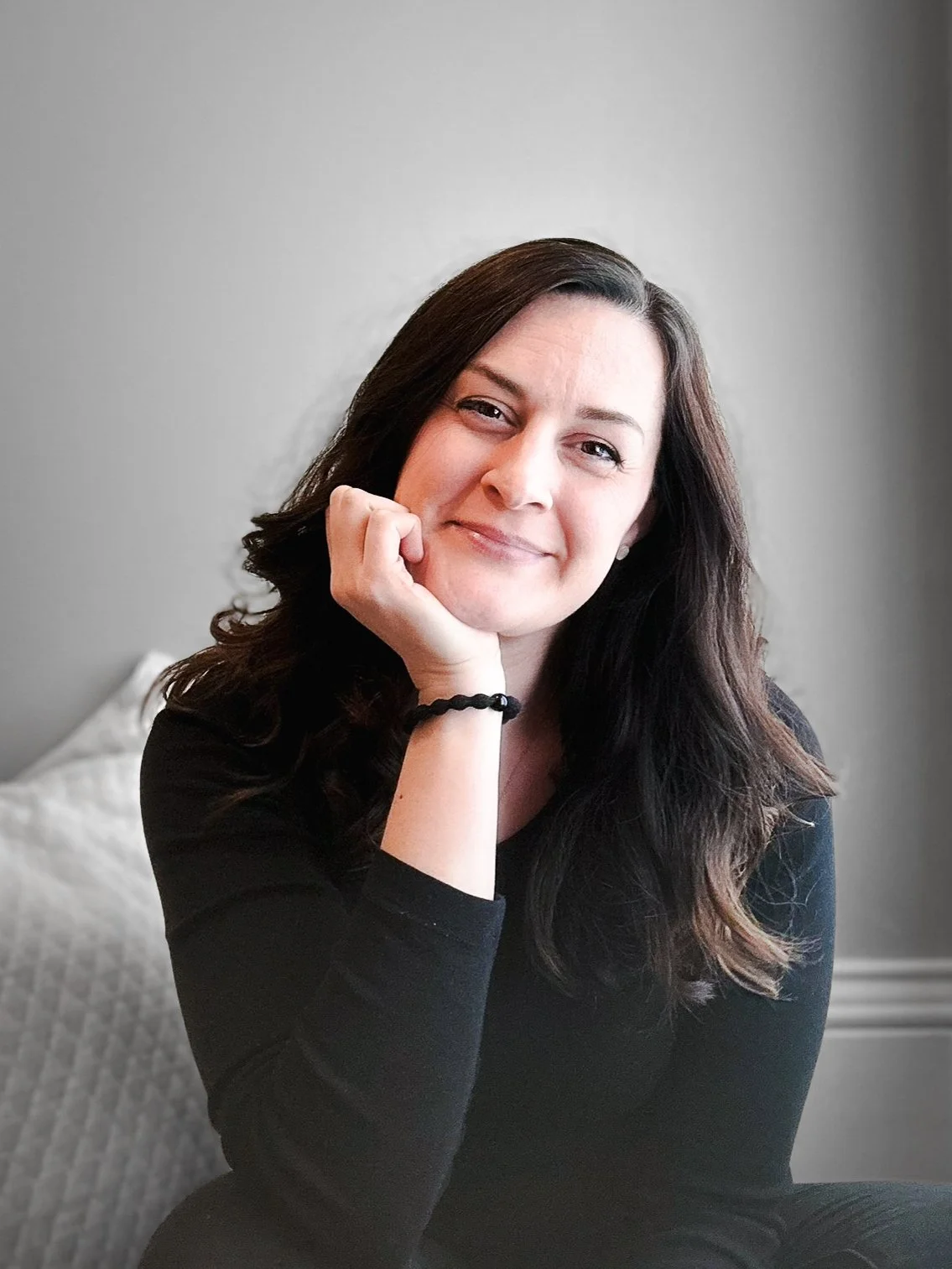 A woman with long dark hair, smiling gently, resting her face on her hand, sitting on a gray patterned cushion in front of a plain light-colored wall.