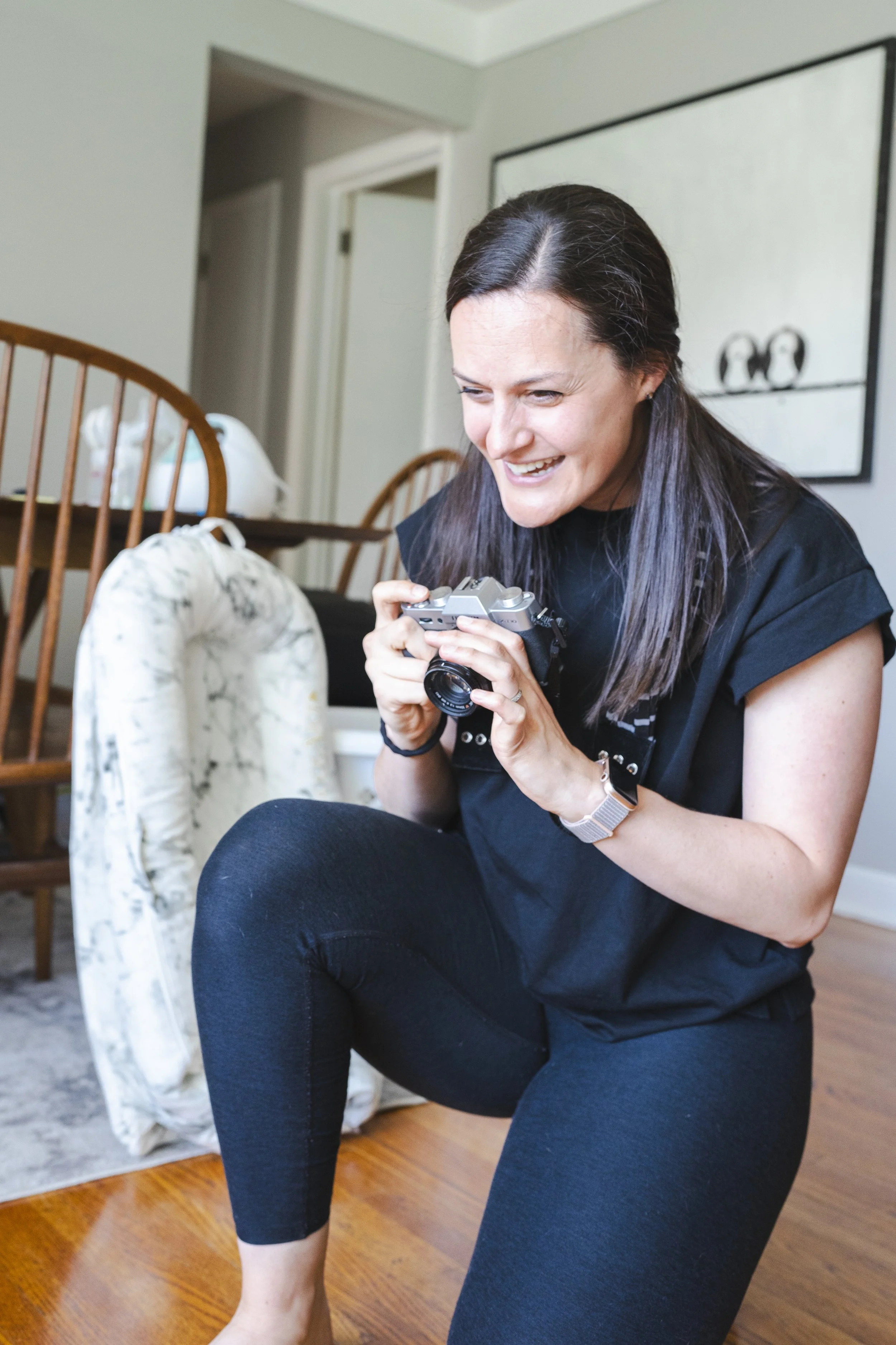 Woman sits on the ground, smiling and holding a camera, in a home interior with wooden floors, a chair, and a wall decorated with a black and white artwork.