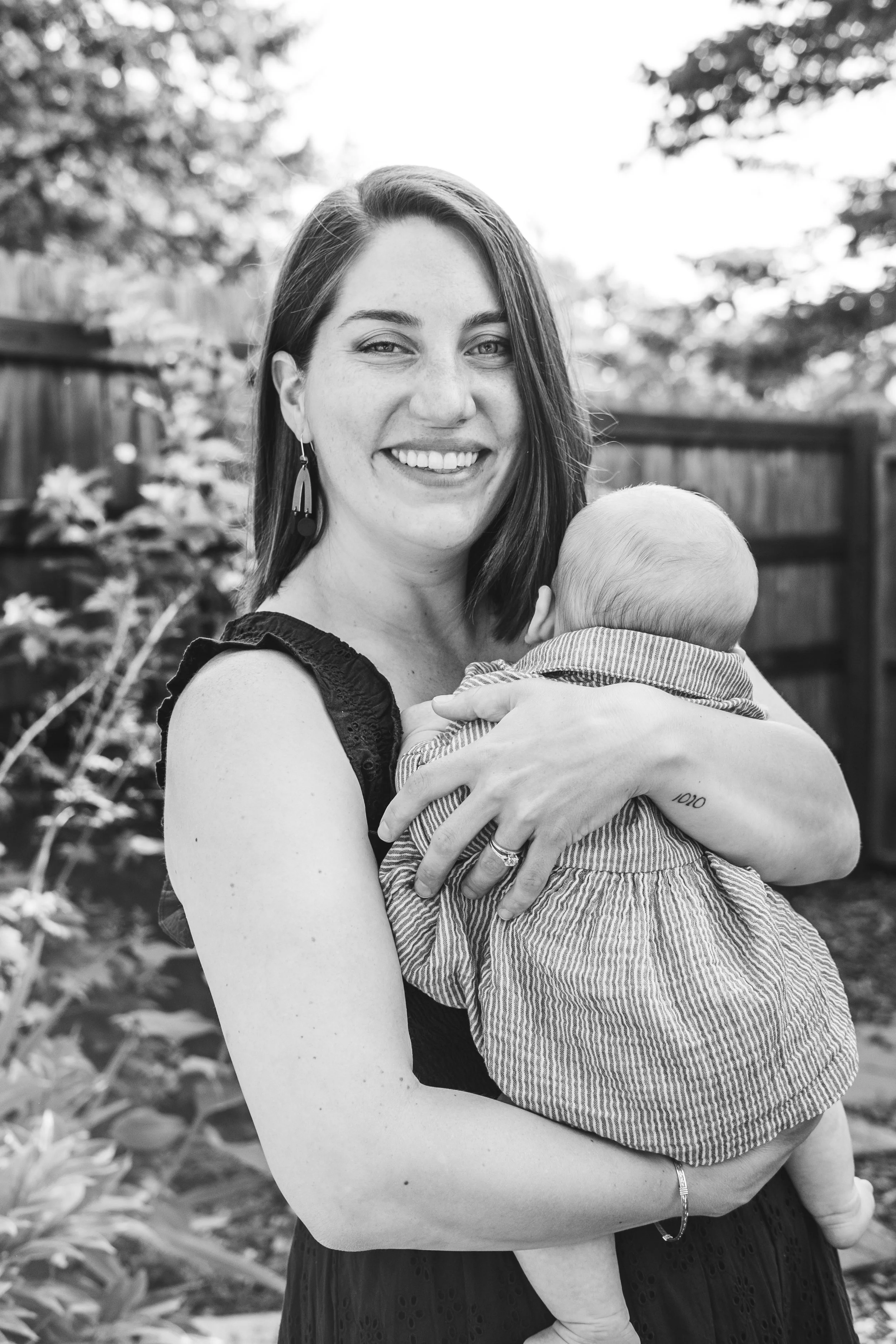 A woman with shoulder-length dark hair and earrings, smiling and holding a baby with a bald head, outdoors with trees and a wooden fence in the background.
