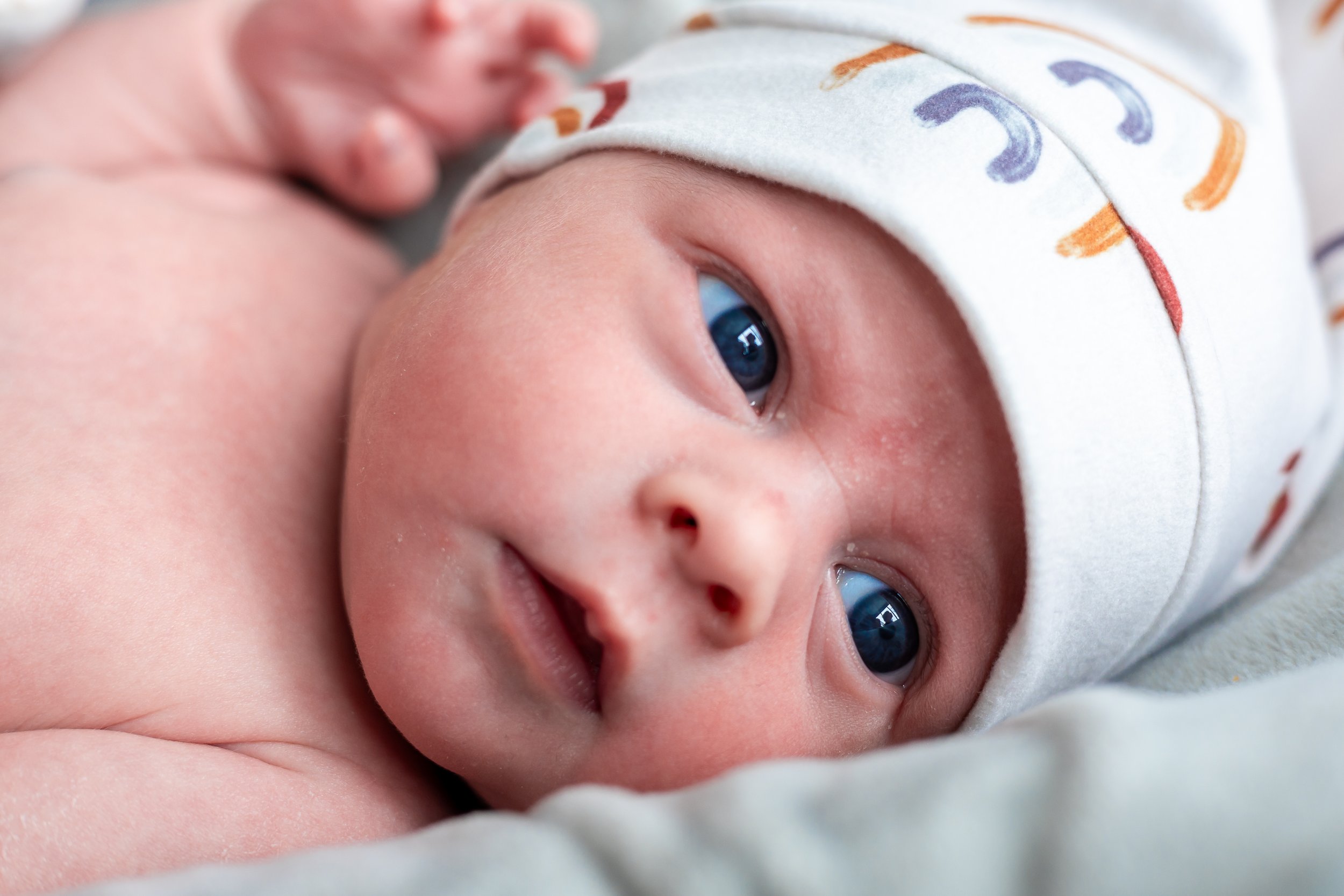 Close-up image of a baby's face with bright blue eyes, wearing a white hat with colorful patterns, lying on a soft surface.