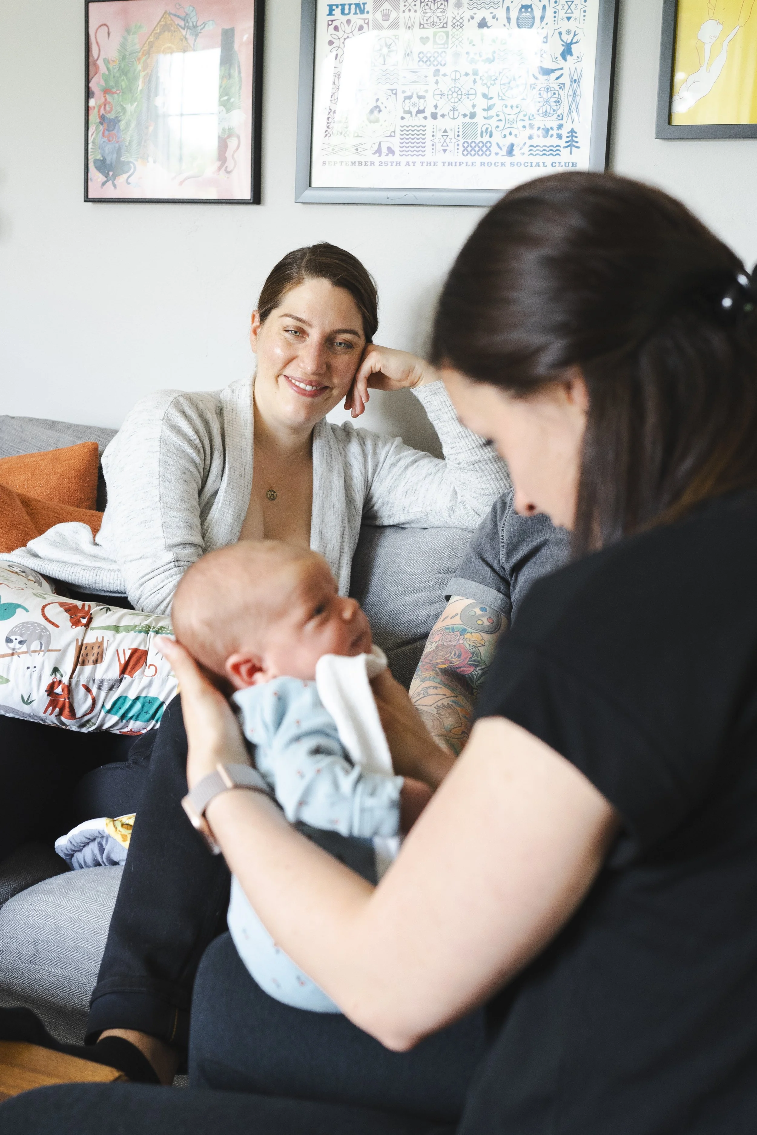 A woman with dark hair in a ponytail is holding a baby dressed in light blue pajamas with colorful animal prints, as another woman with short brown hair and a gray cardigan smiles and leans on her hand, sitting on a gray couch with orange pillows, in a room decorated with framed posters on the wall.