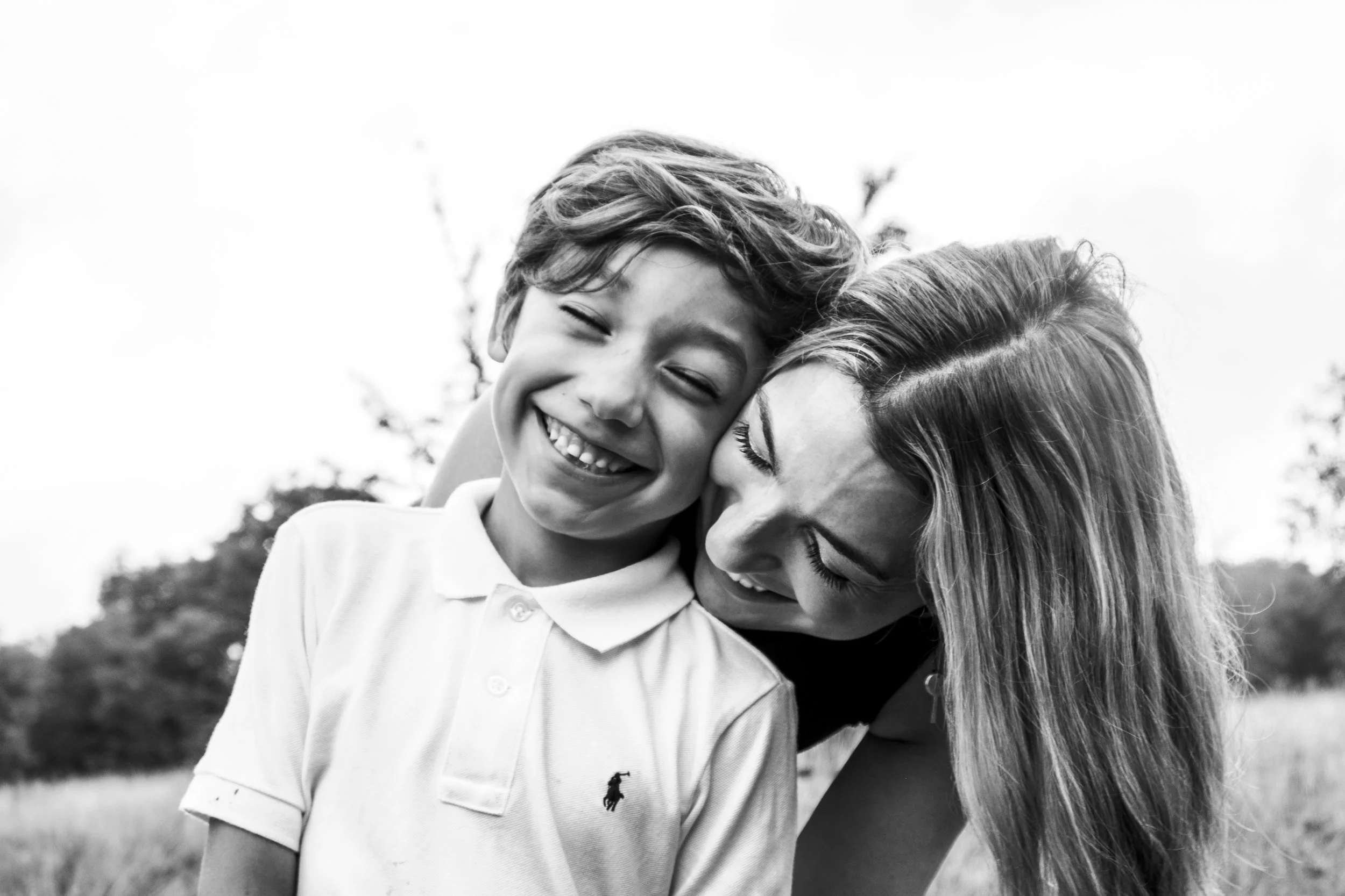 A joyful boy and woman sharing a hug outdoors, smiling with eyes closed, black and white photo.