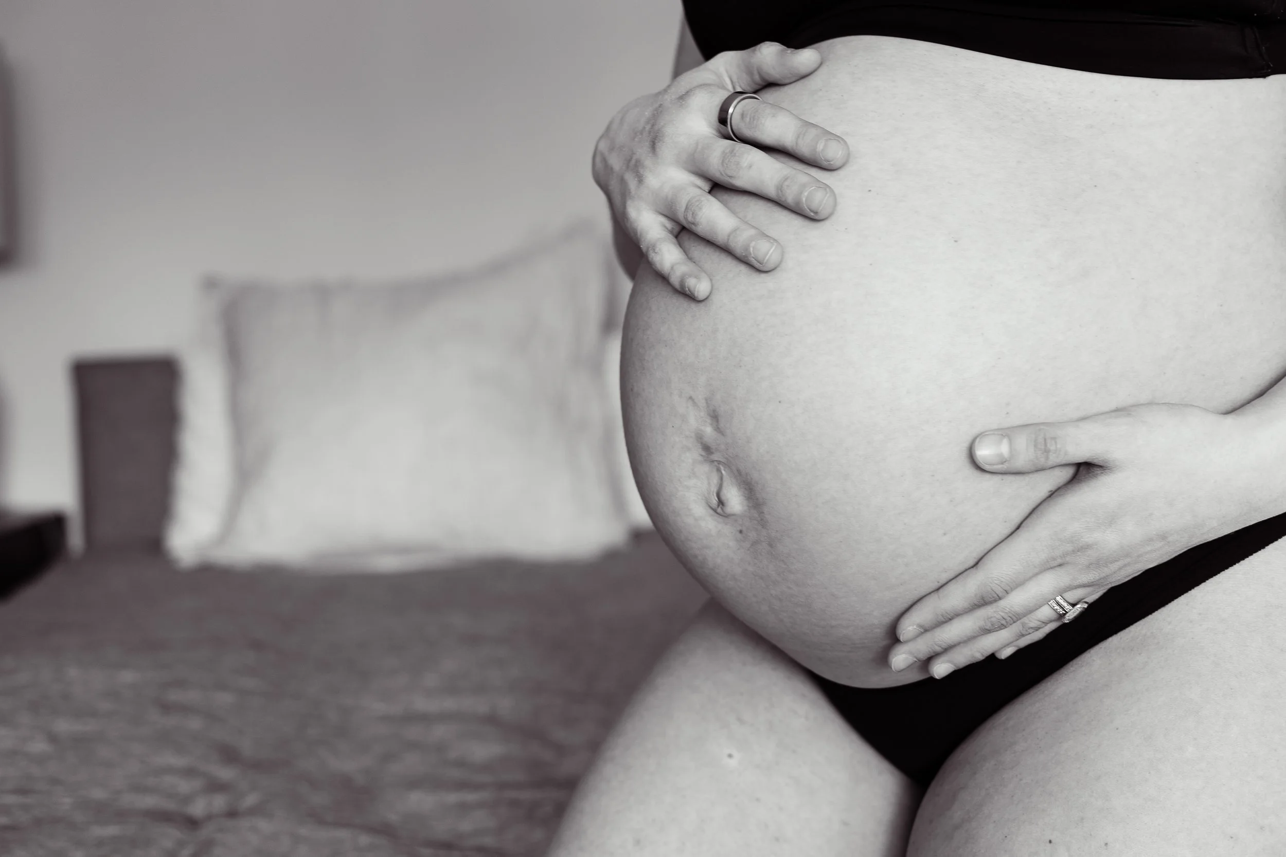 Close-up of a pregnant woman's belly with hands gently resting on it, with one hand wearing a wedding ring, in black and white.