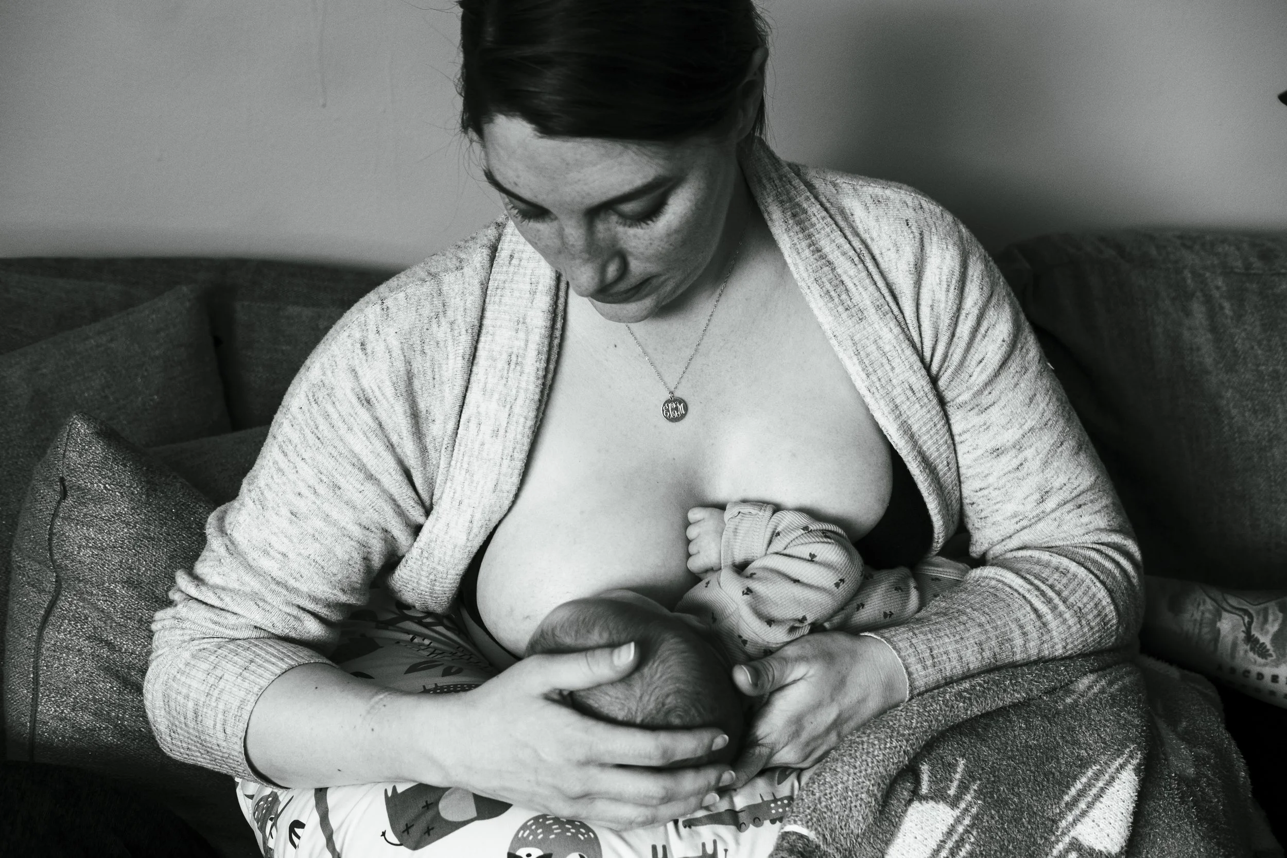A woman breastfeeding a baby while sitting on a couch, with a gentle expression, wearing a necklace, and a cardigan over her shoulders.