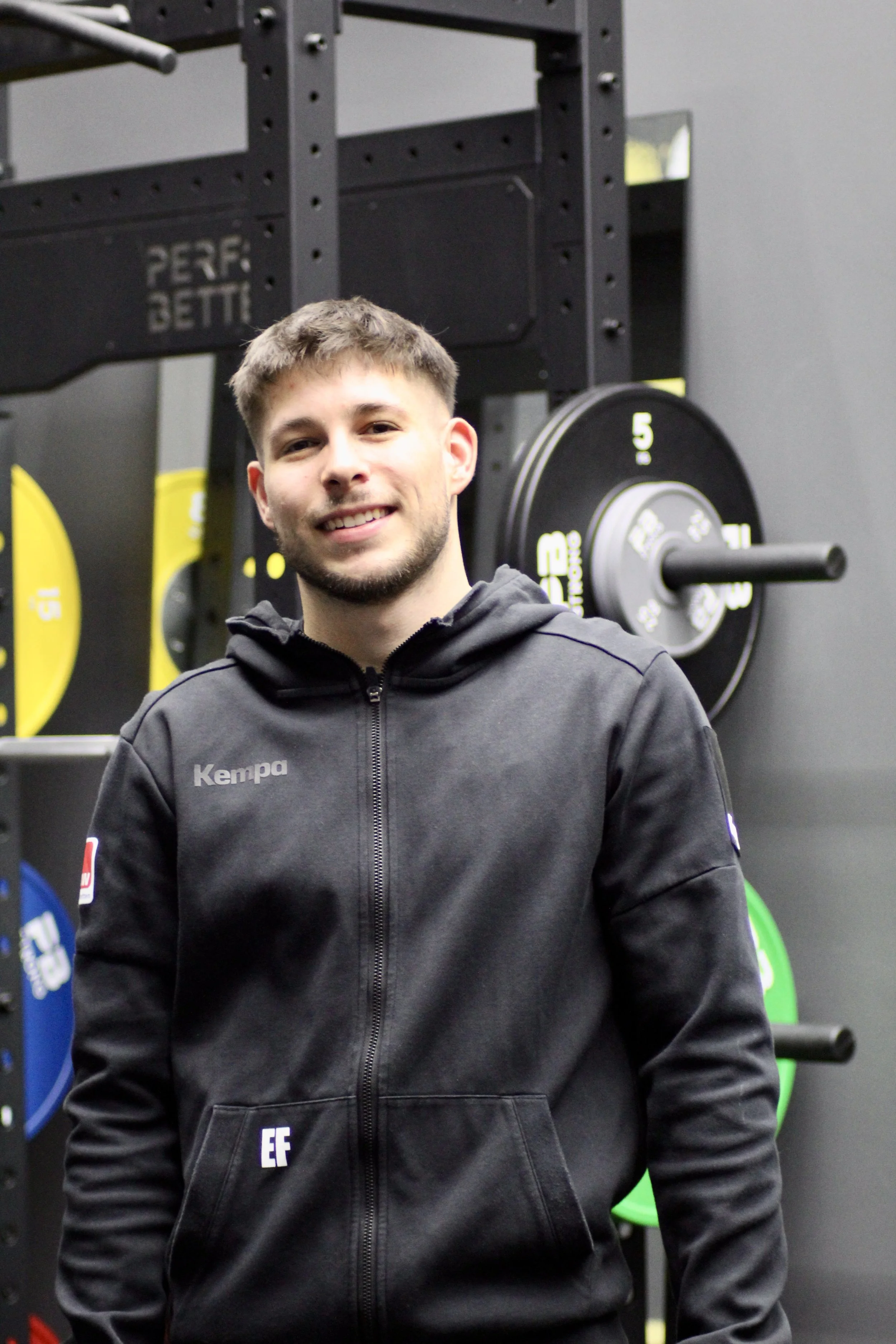 Junge Mann in dunkler Trainingsjacke vor einer Wand mit Gewichtsscheiben, lächelnd.