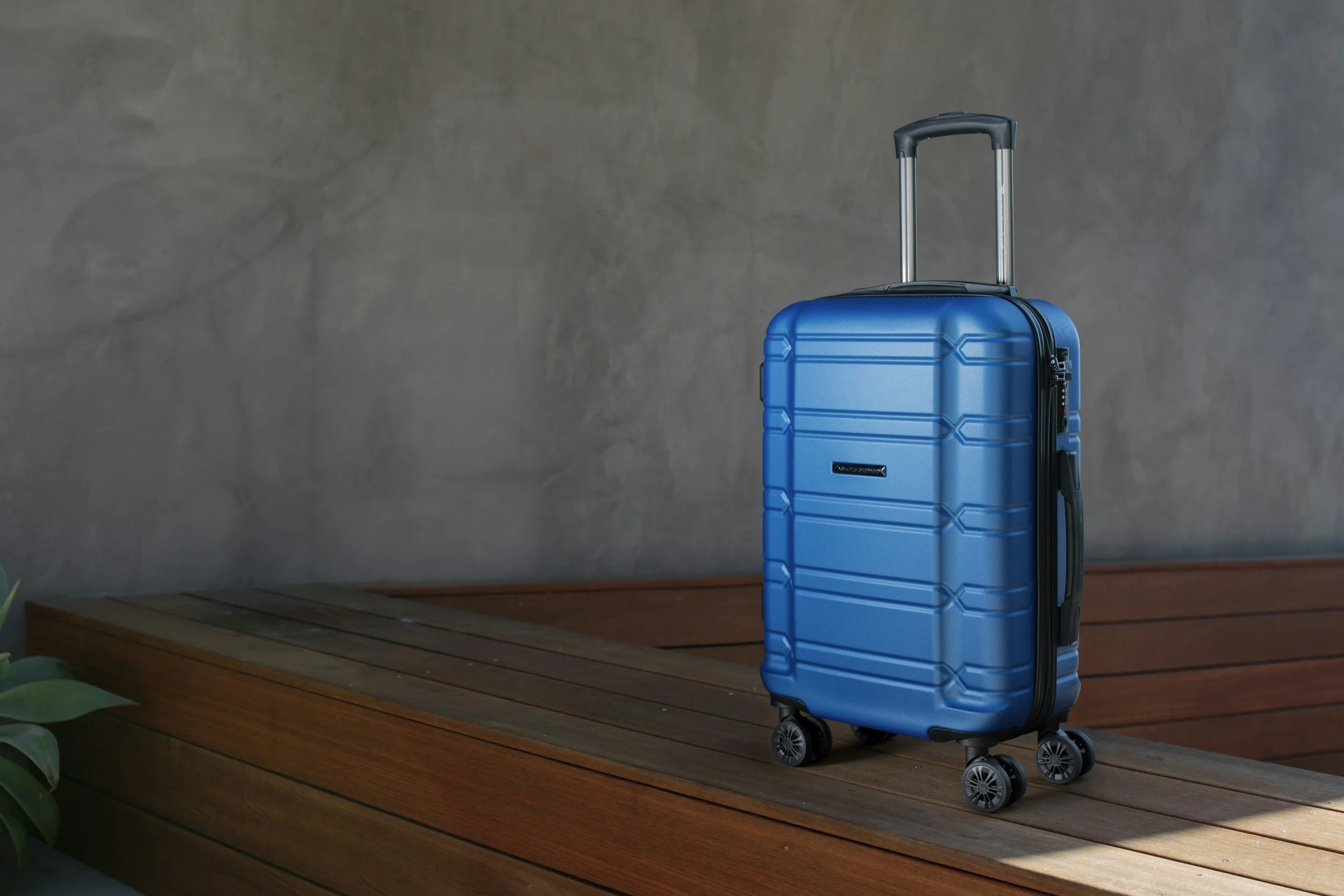 A blue suitcase with a retractable handle and four wheels placed on a wooden surface against a gray wall.