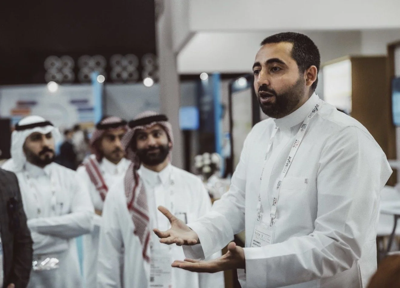 Man in white shirt speaking to a group of men wearing traditional Middle Eastern attire at an indoor event.