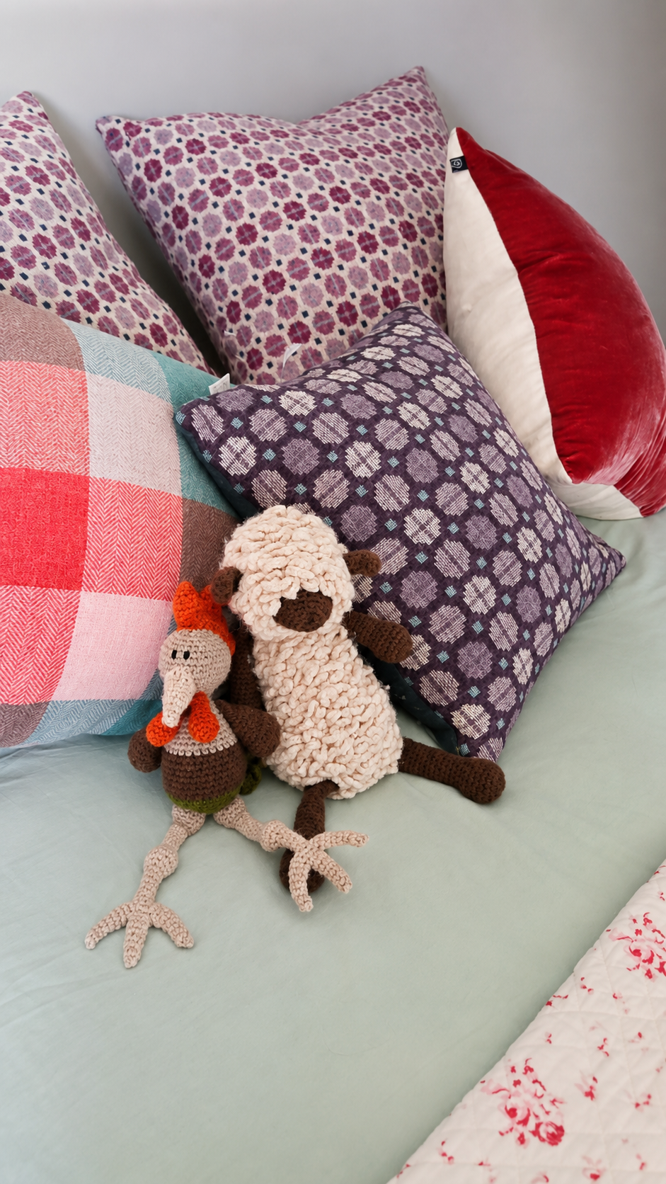 A bed with various pillows and two stuffed animal toys, a crochet chicken and a sheep, resting on a pastel-colored bedsheet. The pillows have different patterns including floral, plaid, and solid colors.