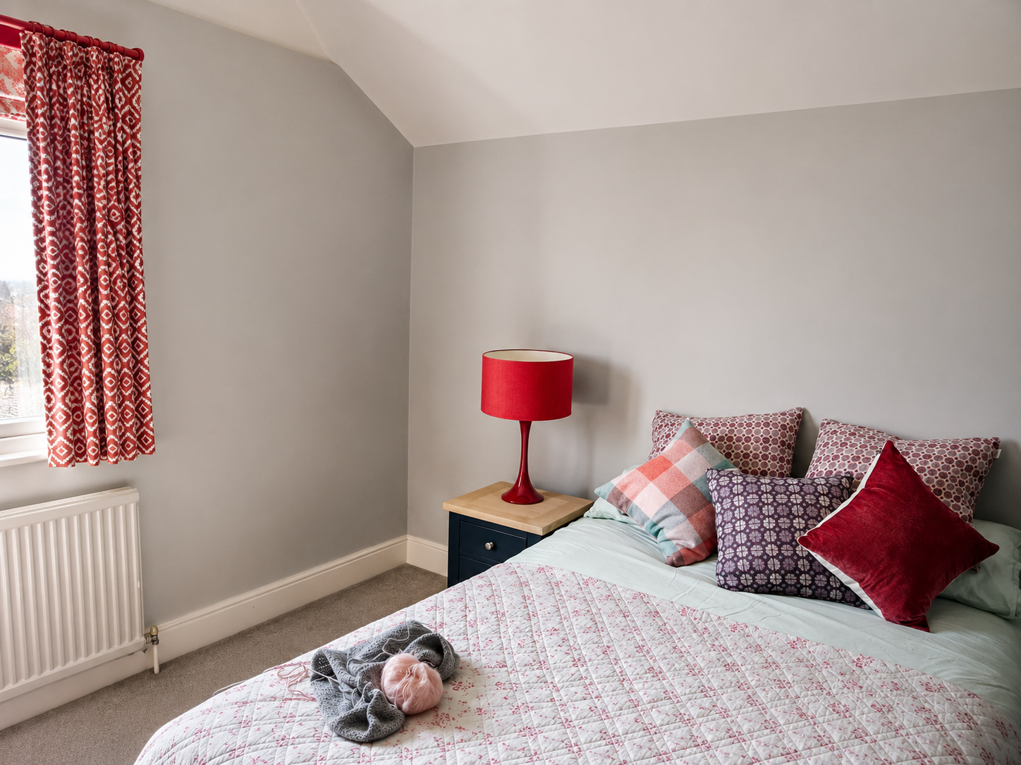 A cozy bedroom with a bed, multiple patterned pillows, and a pink and gray quilt. A pink lamp is on a dark blue nightstand, and a window with a pink and white patterned curtain is on the wall. A small pink stuffed animal and a gray blanket are on the