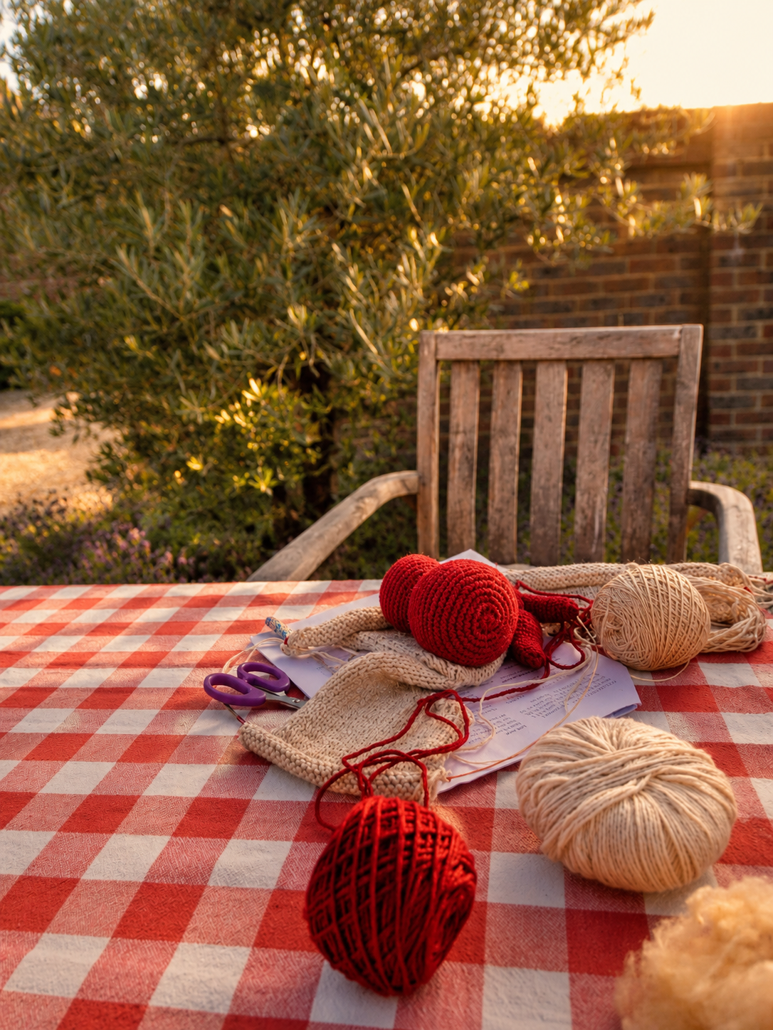 Outdoor table with knitting supplies, including balls of red, beige, and white yarn, knitting needles, and scissors, set on a red and white checkered tablecloth. A wooden chair and greenery are visible in the background, illuminated by warm sunset lighting.