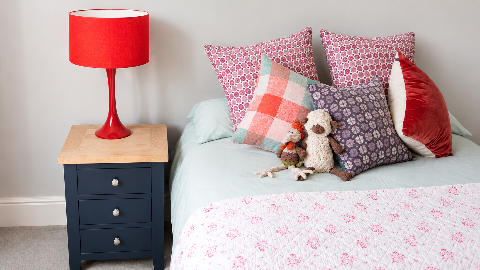 A bedroom with a blue nightstand, red lamp, and a bed with multiple pink, purple, and patterned pillows. There are two small stuffed animals on the bed, one resembling a sheep and the other a monkey, with a white quilted blanket with pink floral embr