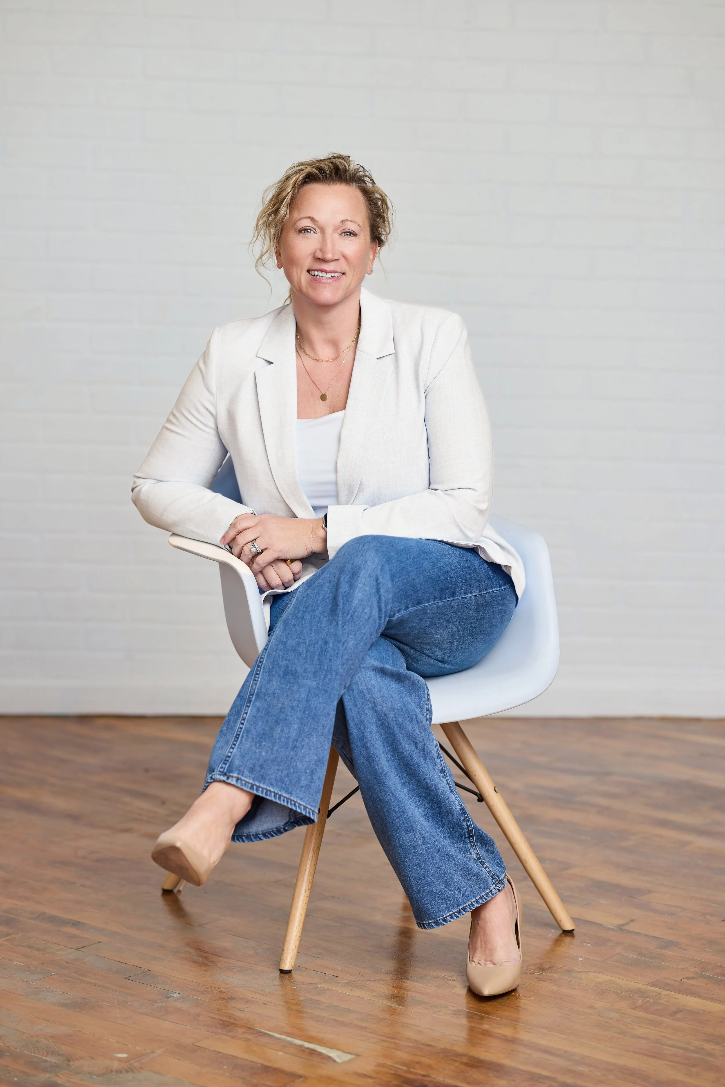 A woman with blonde curly hair sitting on a white chair with wooden legs against a plain white wall. She is smiling, wearing a white blazer, white top, blue jeans, and nude heels. She has layered necklaces and a ring on her left hand. One leg crossed over the other, and her right arm resting on her lap.