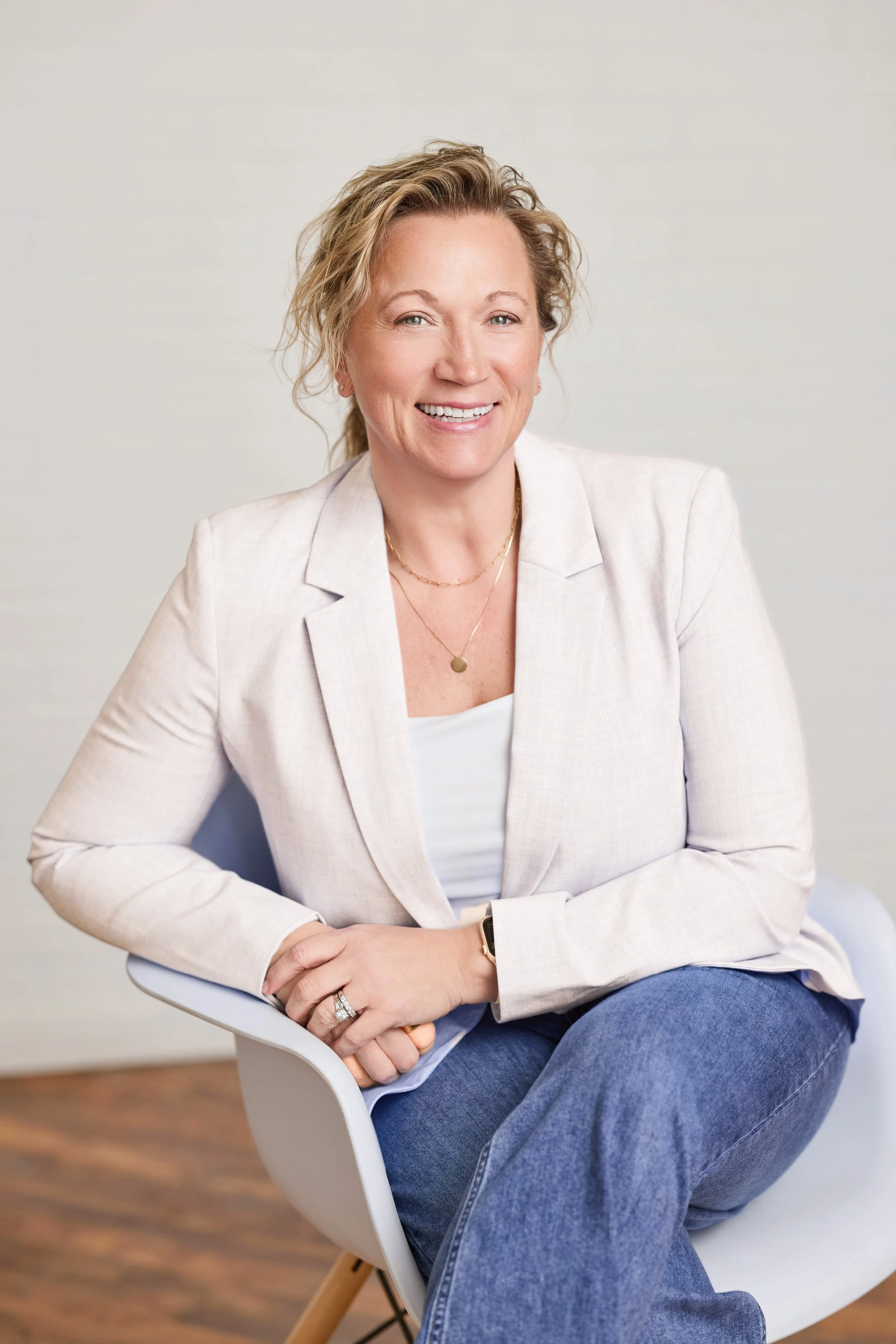 A woman with blonde, curly hair smiling, sitting in a white modern chair, wearing a light beige blazer, white top, and blue jeans, with a plain off-white background.