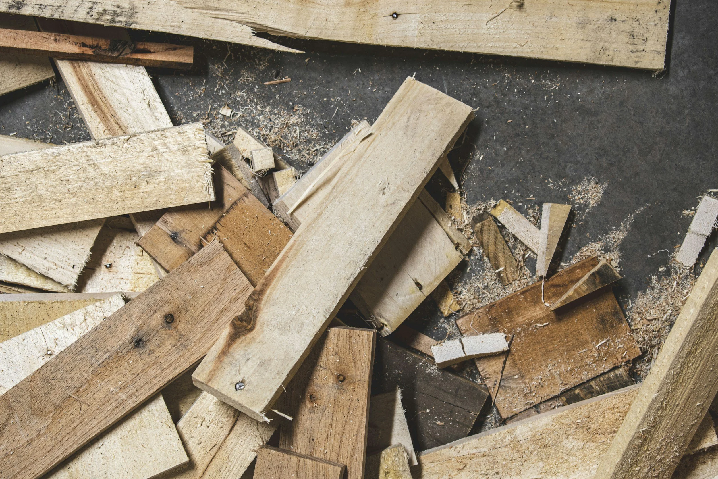 A pile of broken and cut wood pieces on a dark concrete floor with wood shavings and dust.