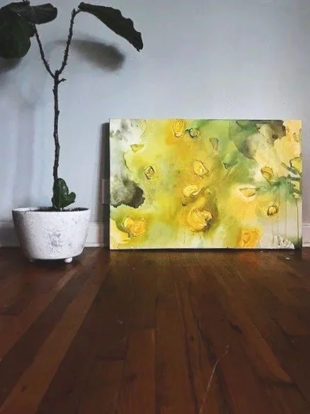 A potted plant with large leaves and a long stem beside a yellow and green abstract painting on a wooden floor.