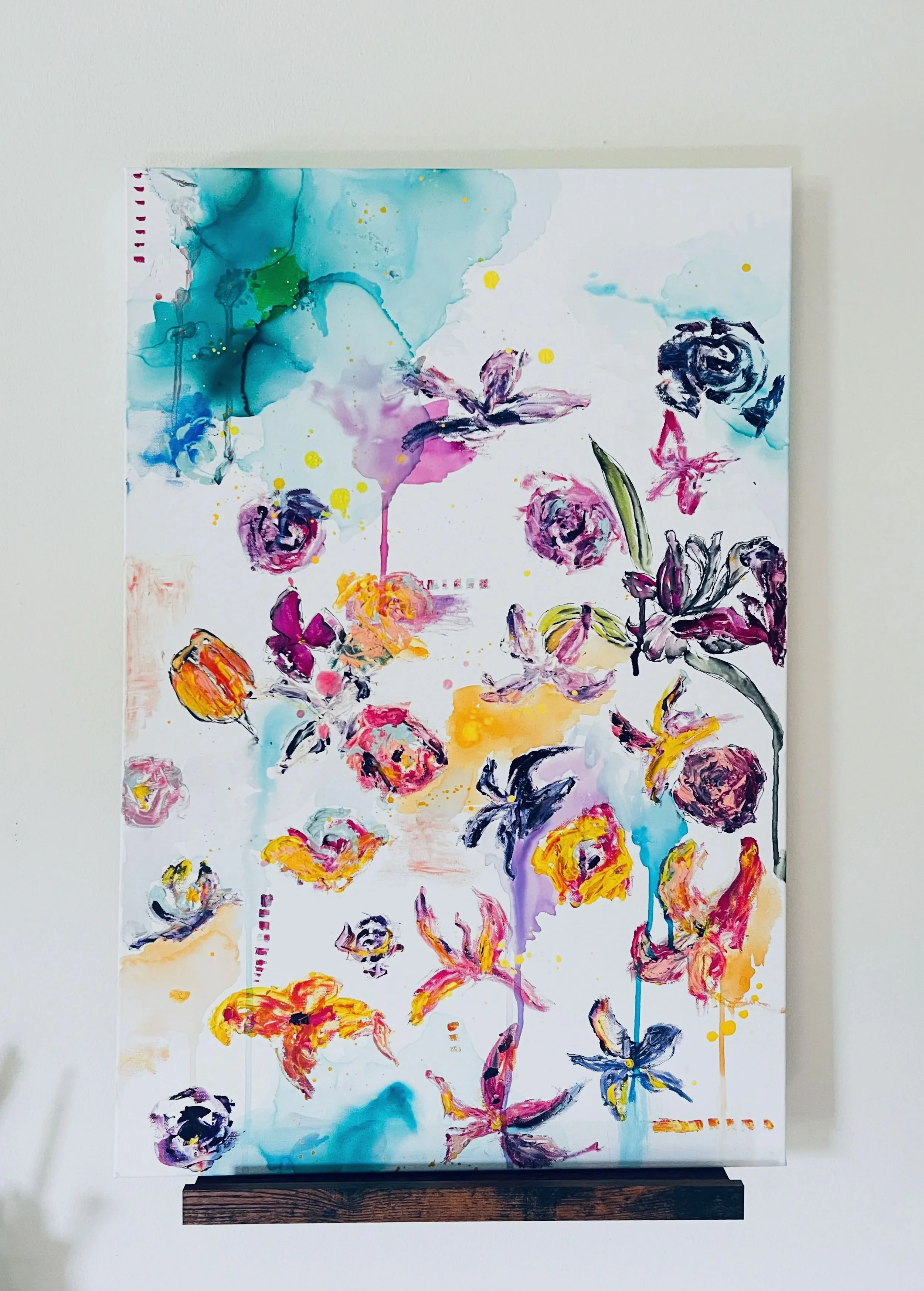 A colorful abstract watercolor painting featuring flowers, butterflies, and splashes of bright colors like blue, pink, purple, yellow, and orange on a vertical paper, mounted on a wall with a wooden ledge.