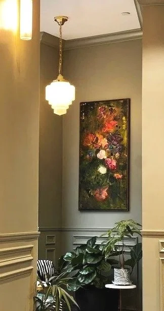 Interior of a room corner with hanging light fixture, framed floral painting on gray wall, and potted plant on side table.