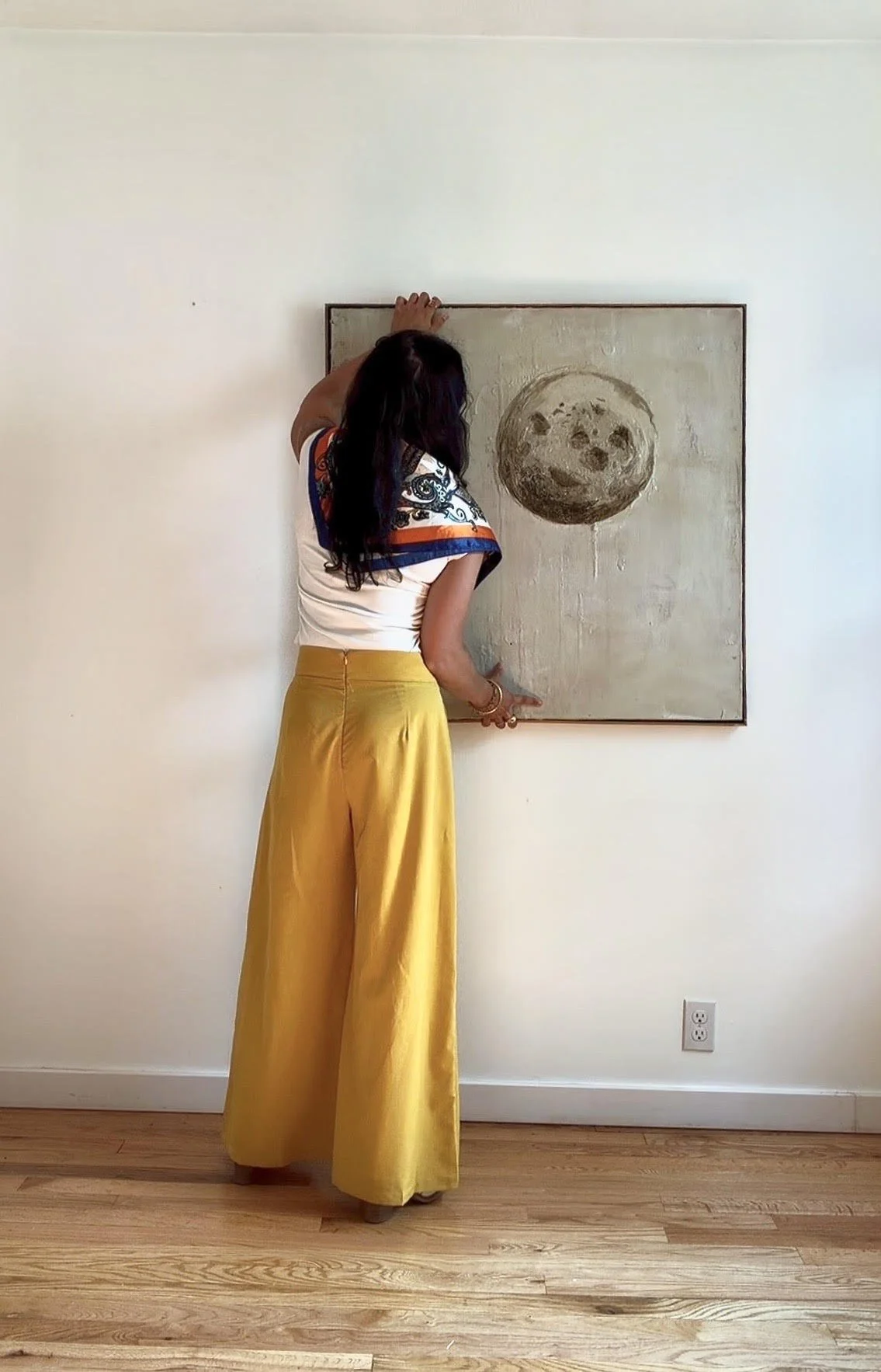 A woman with dark hair, wearing a white top with blue and orange accents and mustard yellow wide-leg pants, stands in front of a white wall with a painting of a smiling moon with a face, touching the top of the painting in an art gallery or room.