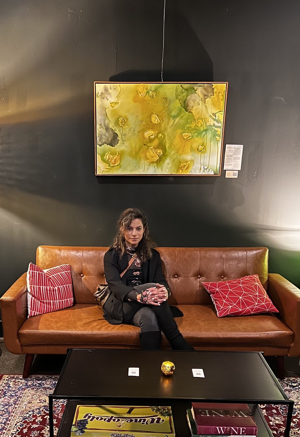 A woman sitting on a brown leather sofa with red and pink patterned cushions in an art gallery. Above her, a framed abstract painting with yellow, green, and gray tones hangs on a black wall, with a small informational placard beside it. A black coffee table with a yellow candle holder and a couple of books is in front of the sofa, and a patterned rug covers the floor.