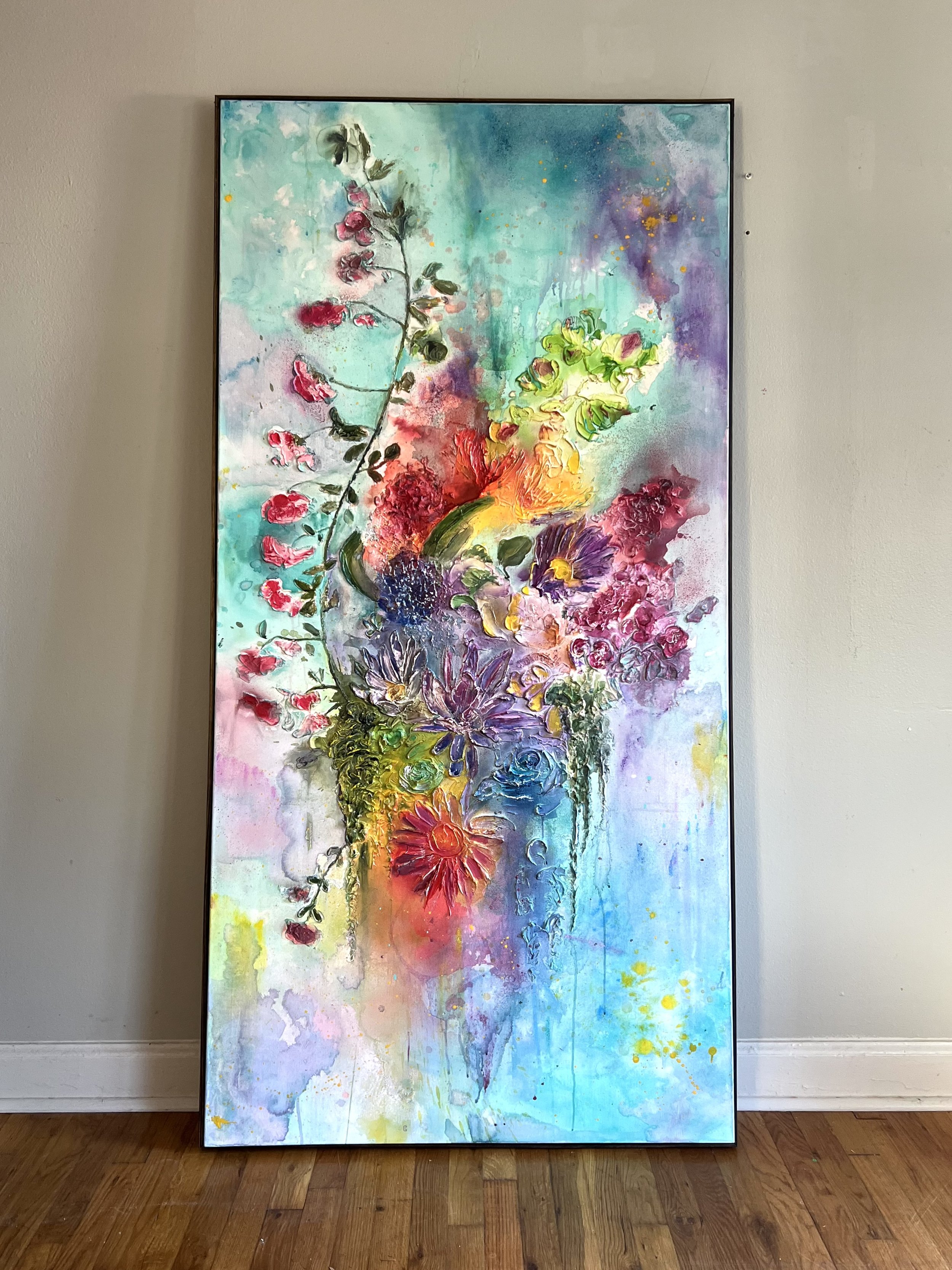 A large, colorful abstract floral painting in a frame, leaning against a beige wall on a wooden floor.
