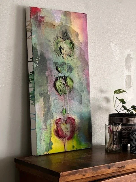 Abstract watercolor painting of three green apples with pink and red accents, on a multicolored background, leaning against a wall on a wooden surface.