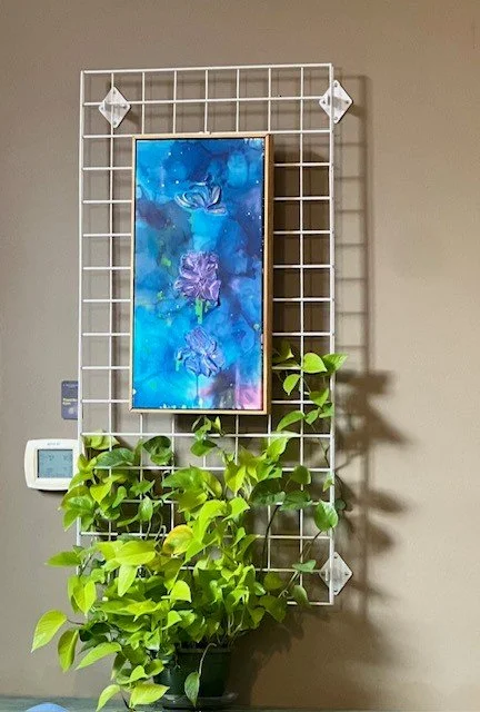 Wall-mounted white grid with a hanging blue and purple floral painting and a green potted plant below it.
