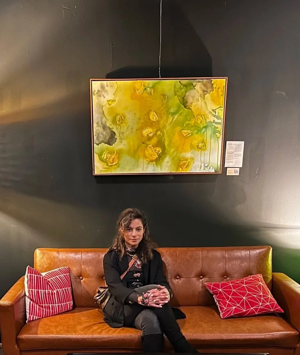 A woman sitting on a brown leather sofa with two red patterned cushions, in front of a black wall with a colorful abstract painting hanging above her.