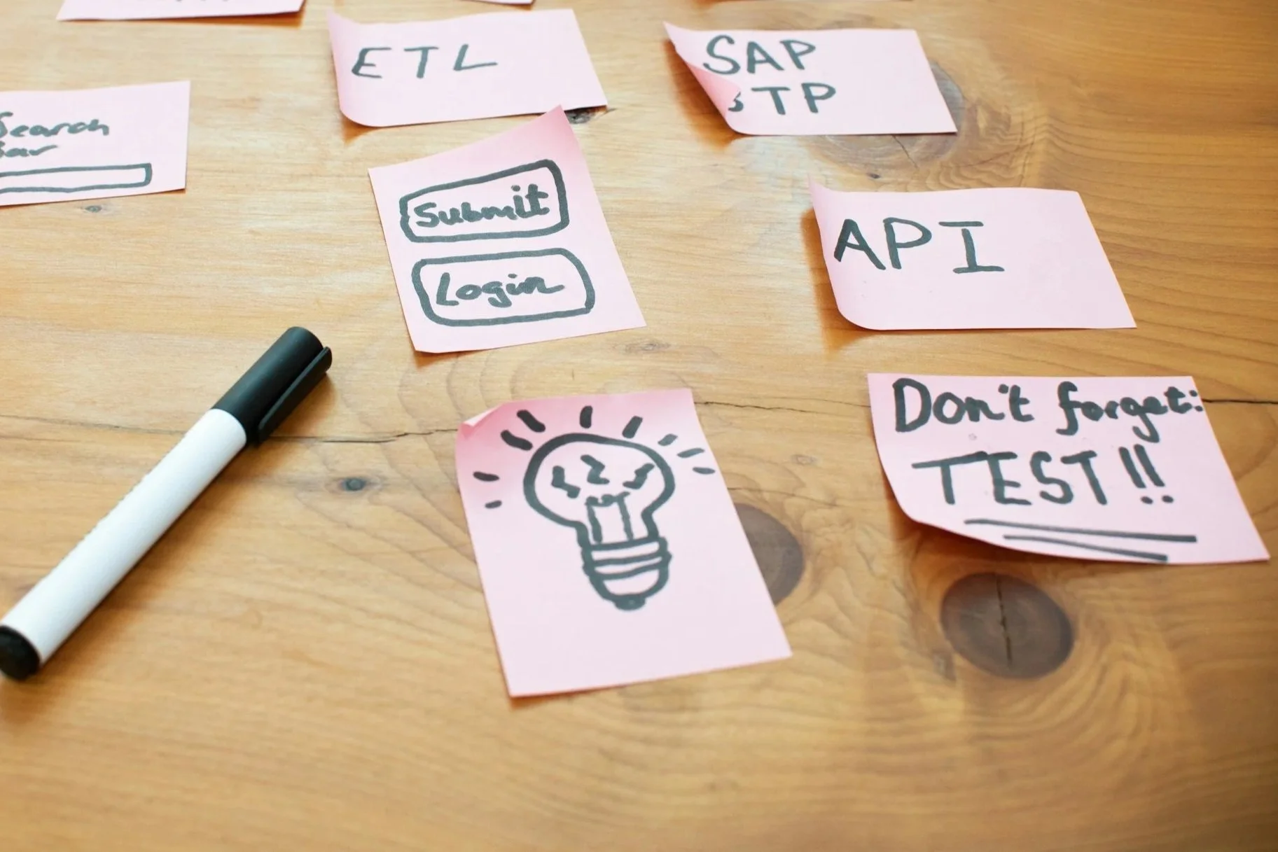 Several pink sticky notes with handwritten text and drawings are scattered on a wooden table. Some notes contain words like 'ETL,' 'SAP,' 'API,' 'STP,' and phrases like 'submit', 'login', 'Don't forget: TEST!!.' There is also a drawing of a lightbulb with lines indicating it is lit, and a black and white marker pen on the left side of the image.