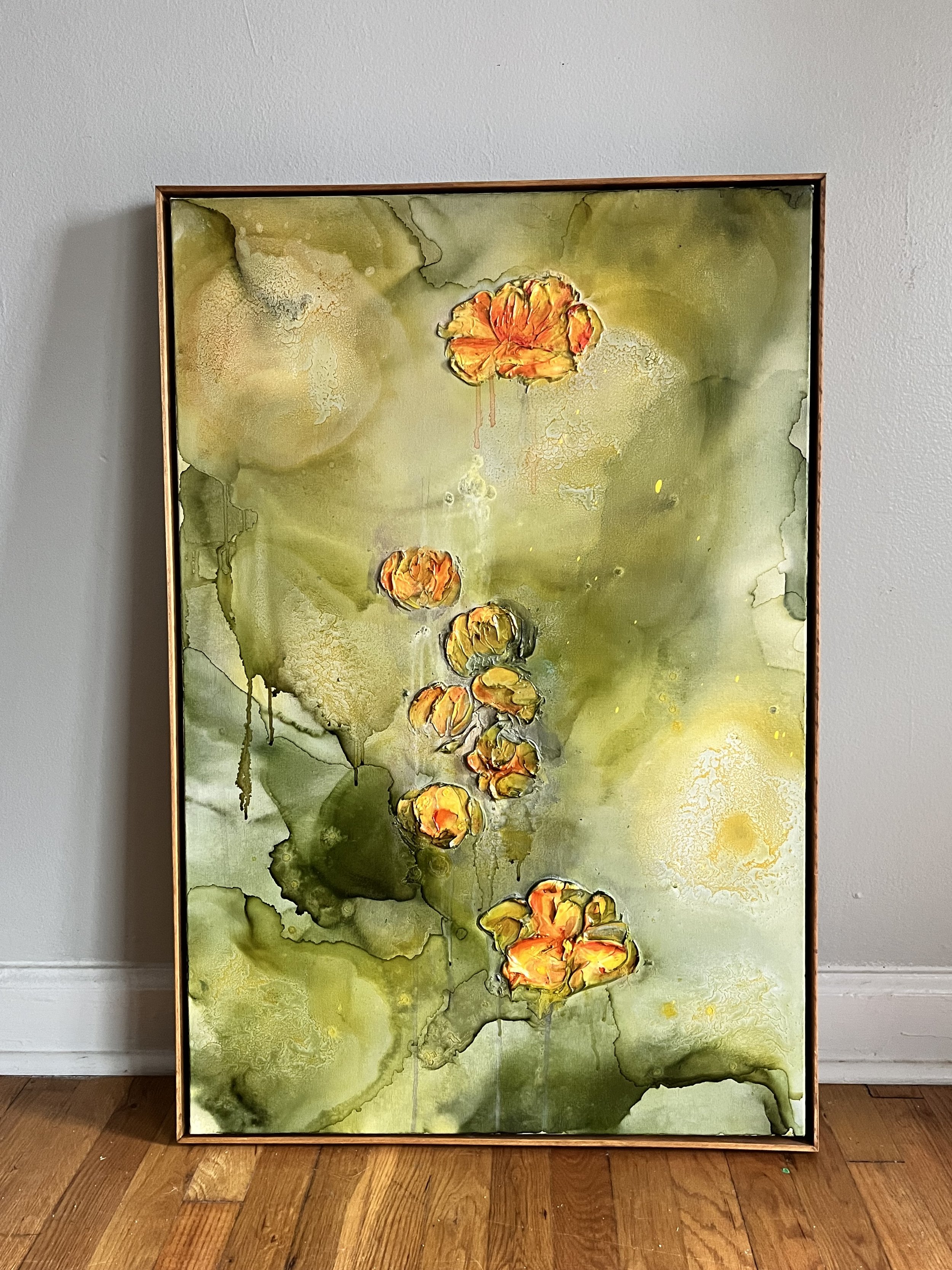 A framed abstract watercolor painting of orange flowers on a green background, leaning against a gray wall with hardwood flooring.
