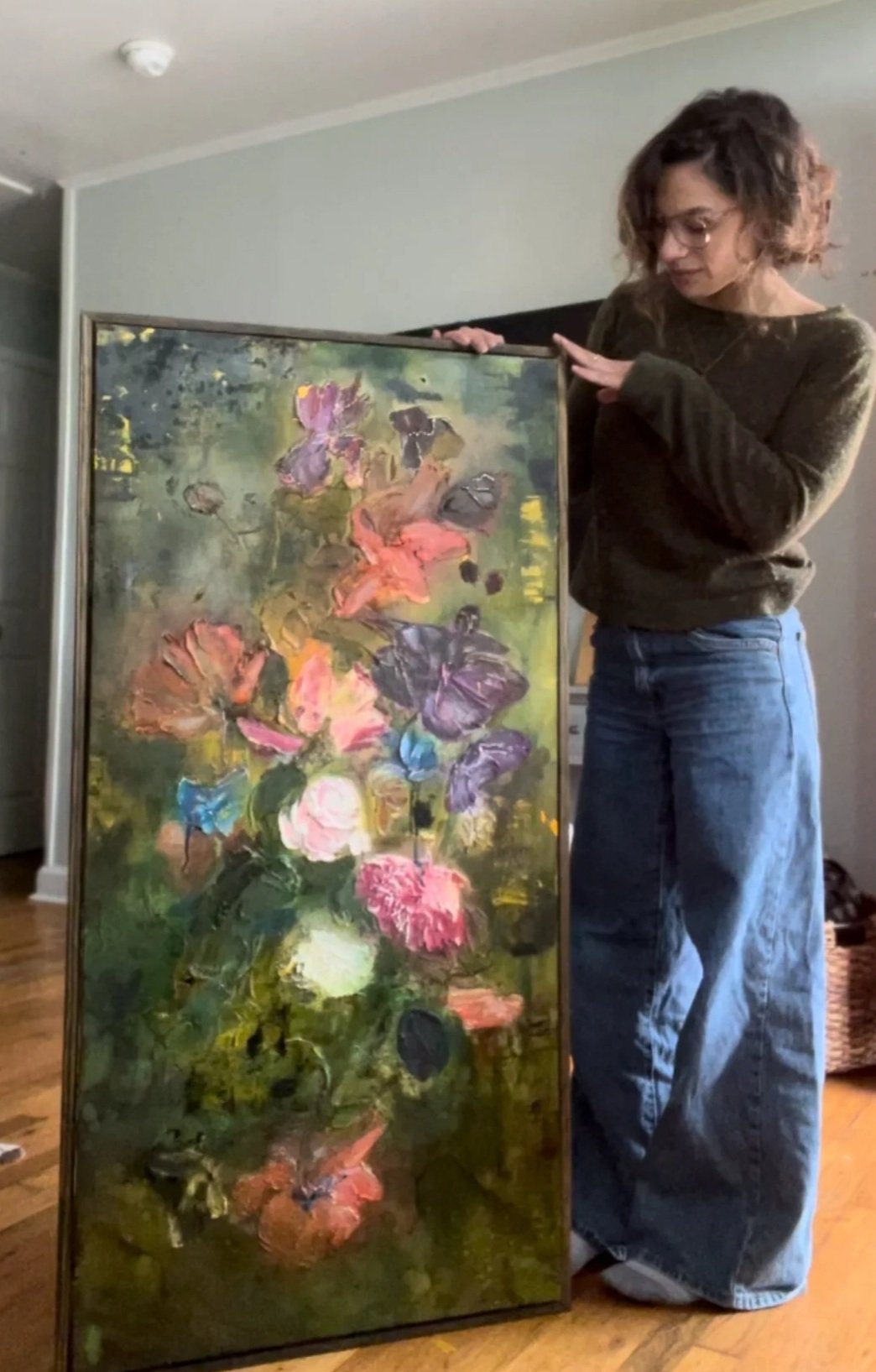 A woman with curly brown hair and glasses, wearing a dark sweater and loose jeans, stands next to an abstract floral painting on a wooden floor in a room with gray walls.